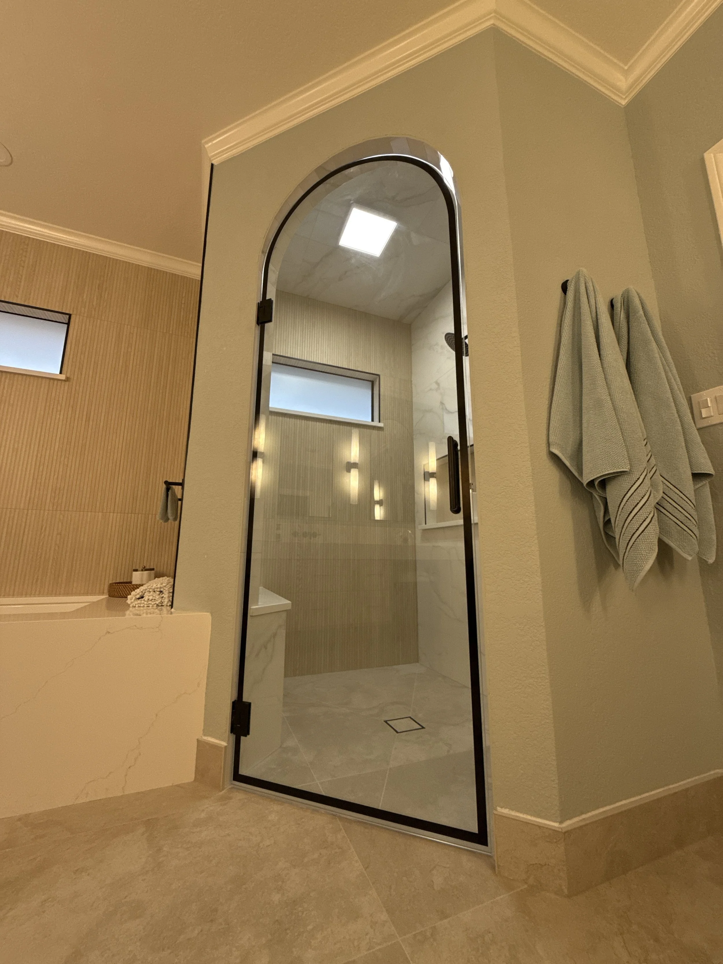 Bathroom with glass shower door, beige tiles, and towels hanging on a wall.