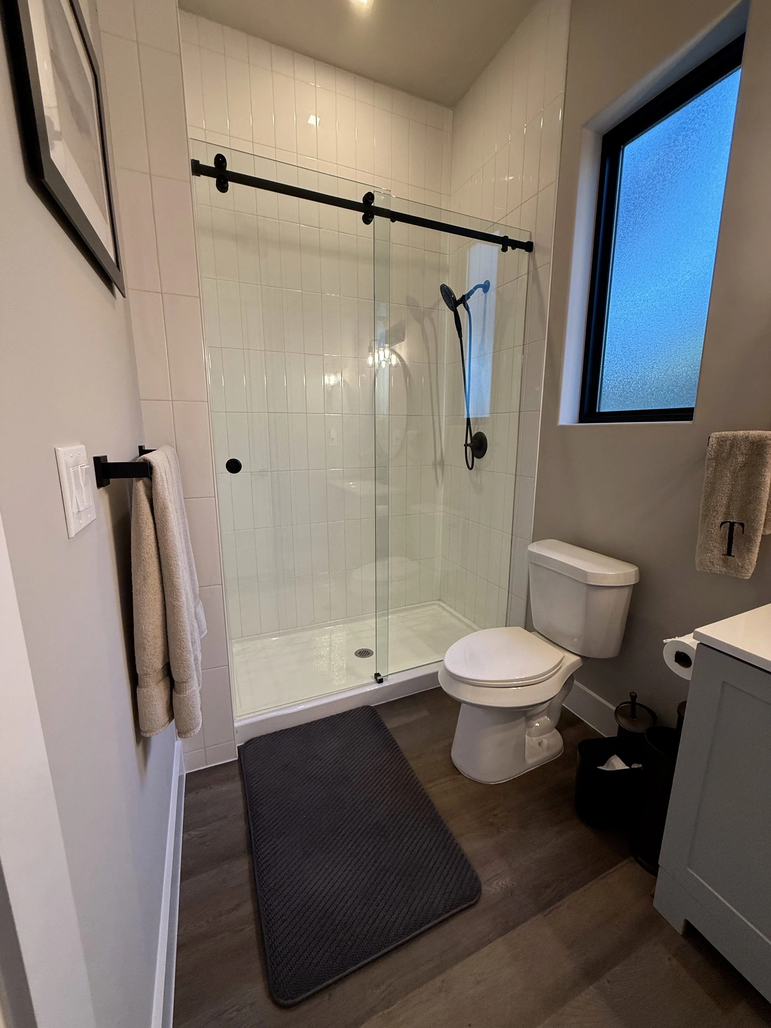 Modern bathroom with a walk-in glass shower, a white toilet, a gray rug, and a window with frosted glass.