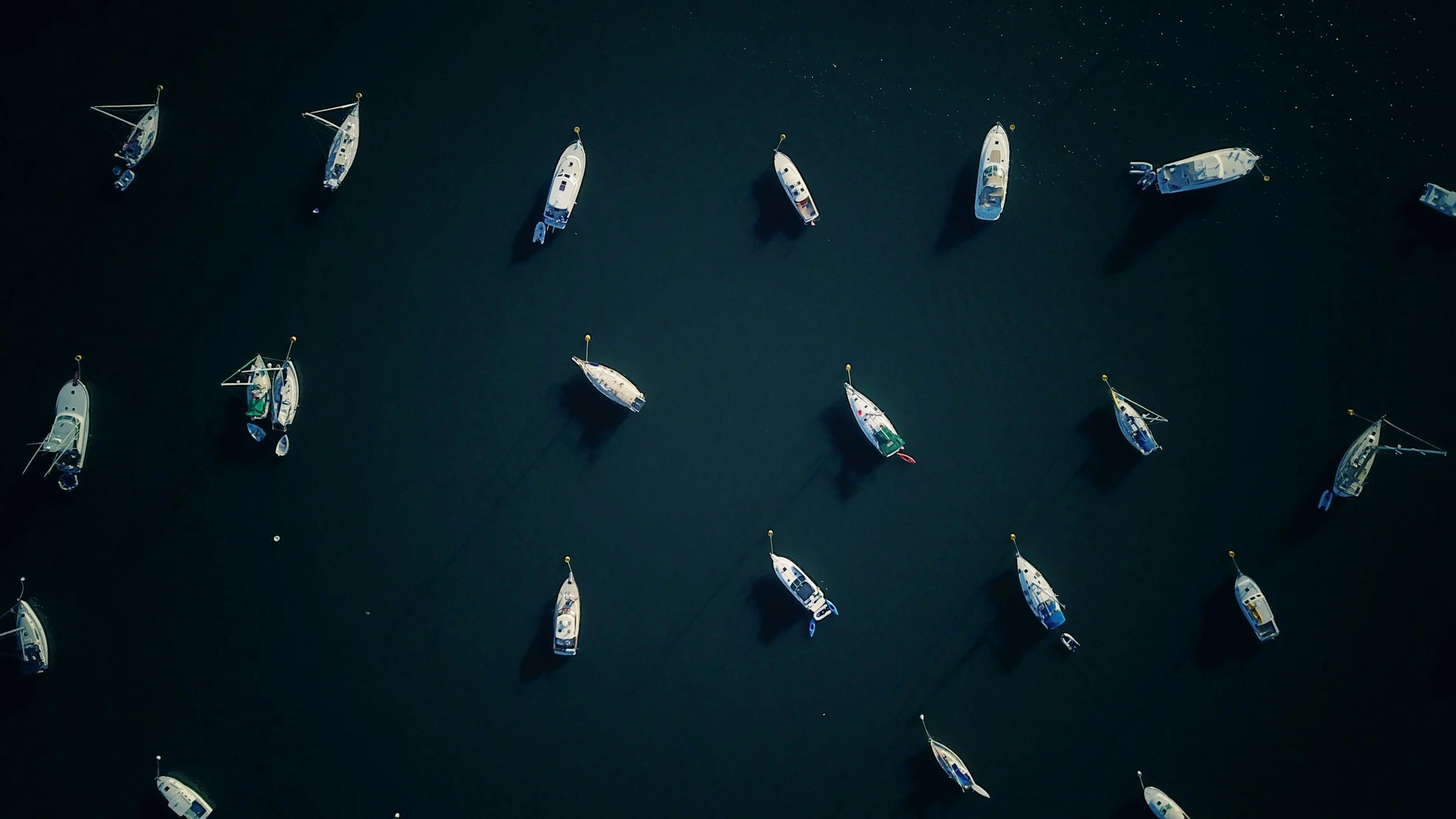 Aerial view of sailboats anchored in dark water, arranged in a pattern with shadows cast on the water.