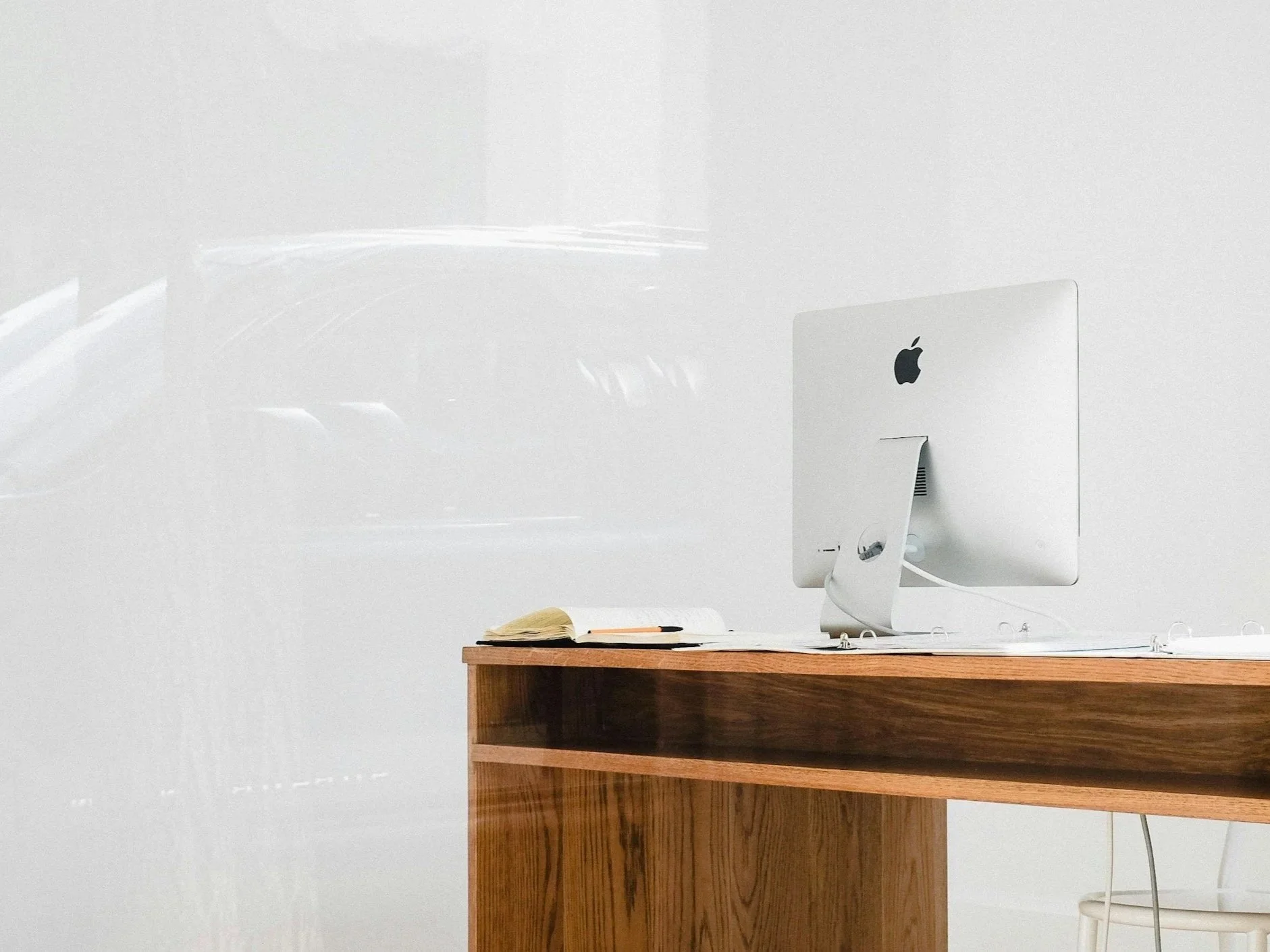 An Apple desktop computer on a wooden desk with a book and a notebook, against a plain white wall. Working on a document translation.