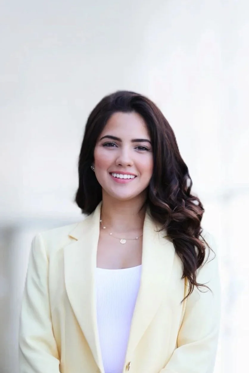 Client review. Portrait of a young woman with dark brown, wavy hair, wearing a light yellow blazer and a white top, standing outdoors with a neutral blurred background, smiling at the camera.