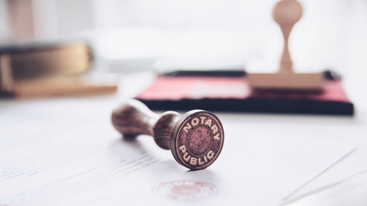 A rubber stamp with the words "Notary Public" on it resting on official documents, with more stamps and office supplies in the blurred background.