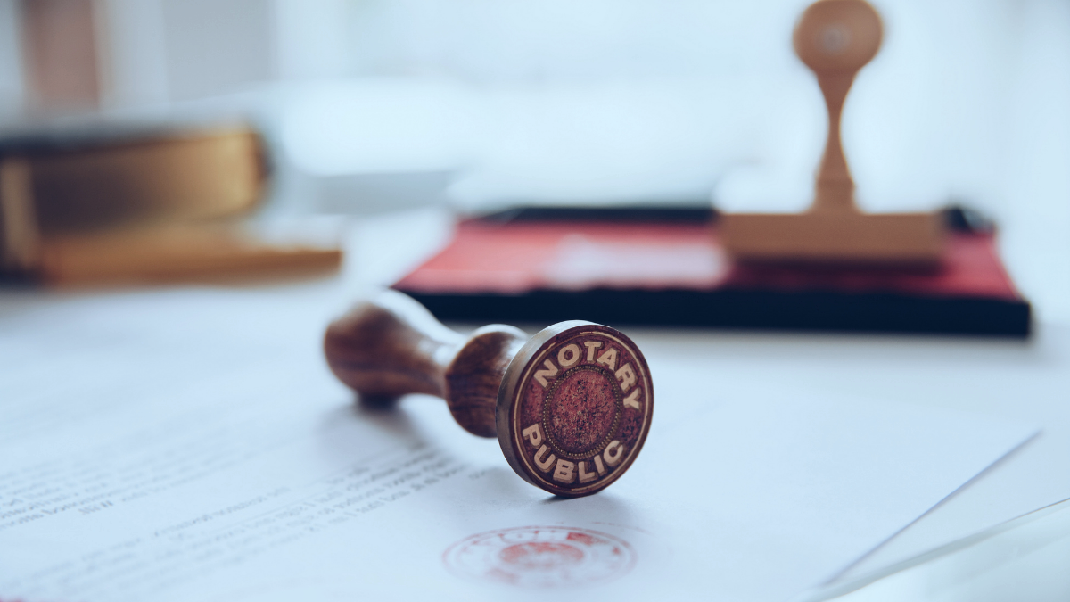 A rubber stamp with the words "Notary Public" stamped on a document with a red ink stamp and other official stamps or seals, and blurred background including a red and black stamp pad and other office items.