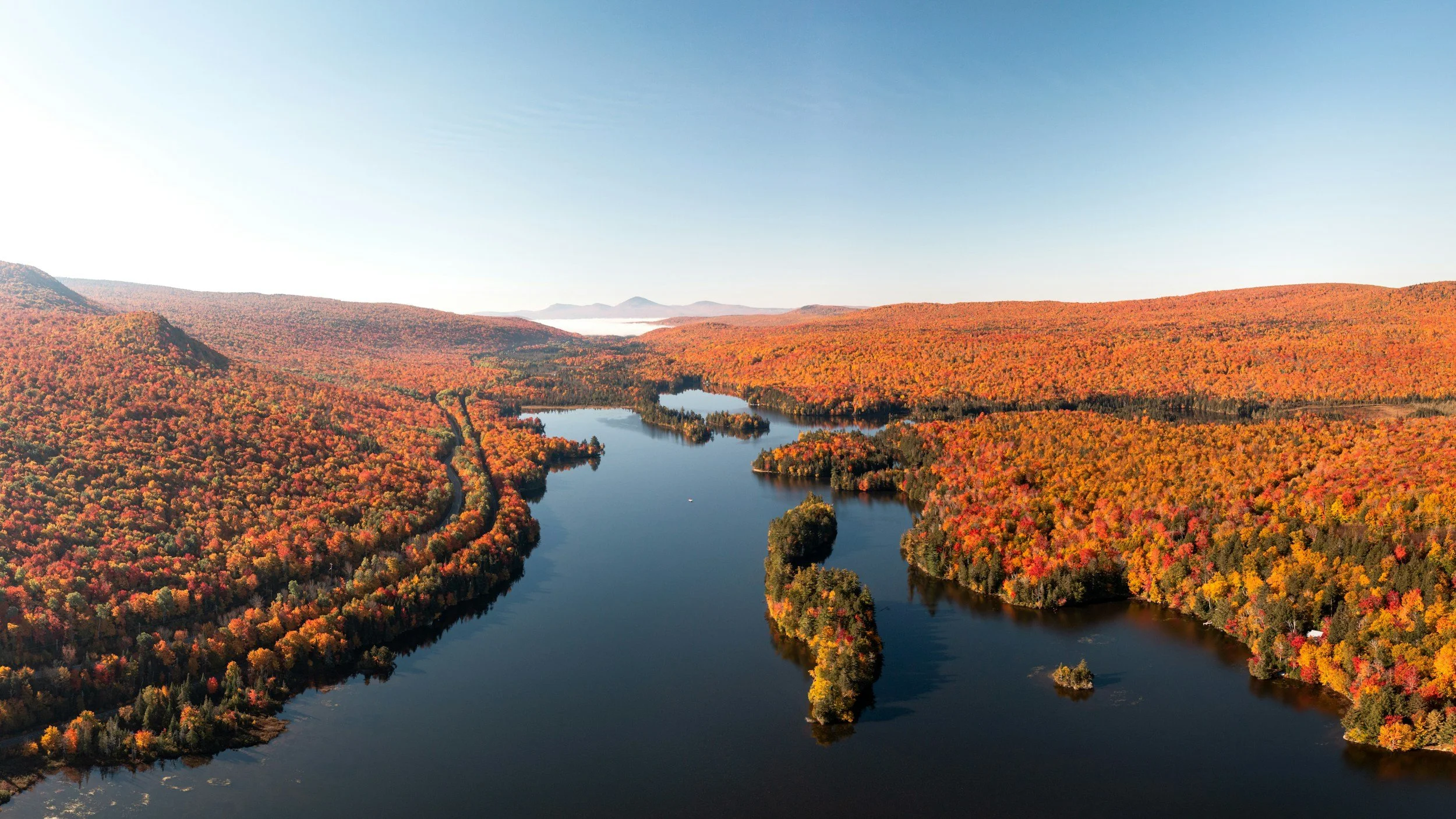 Aerial view of a river winding through a forested landscape with autumn foliage in red, orange, and yellow hues, surrounded by rolling hills under a clear blue sky.