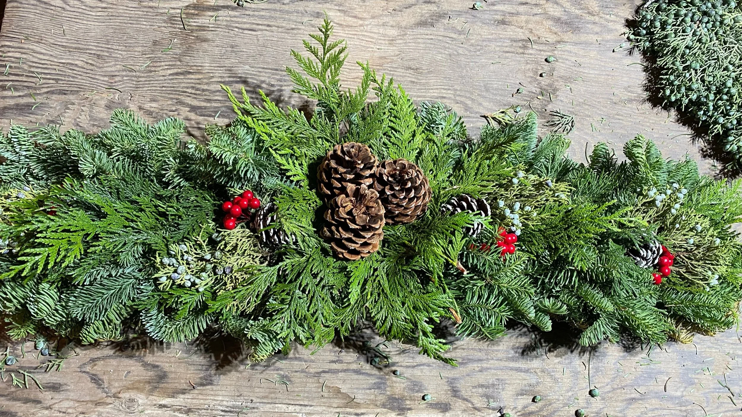 A holiday decoration made of green evergreen branches, pinecones, red berries, and small silver decorations, arranged on a wooden surface.