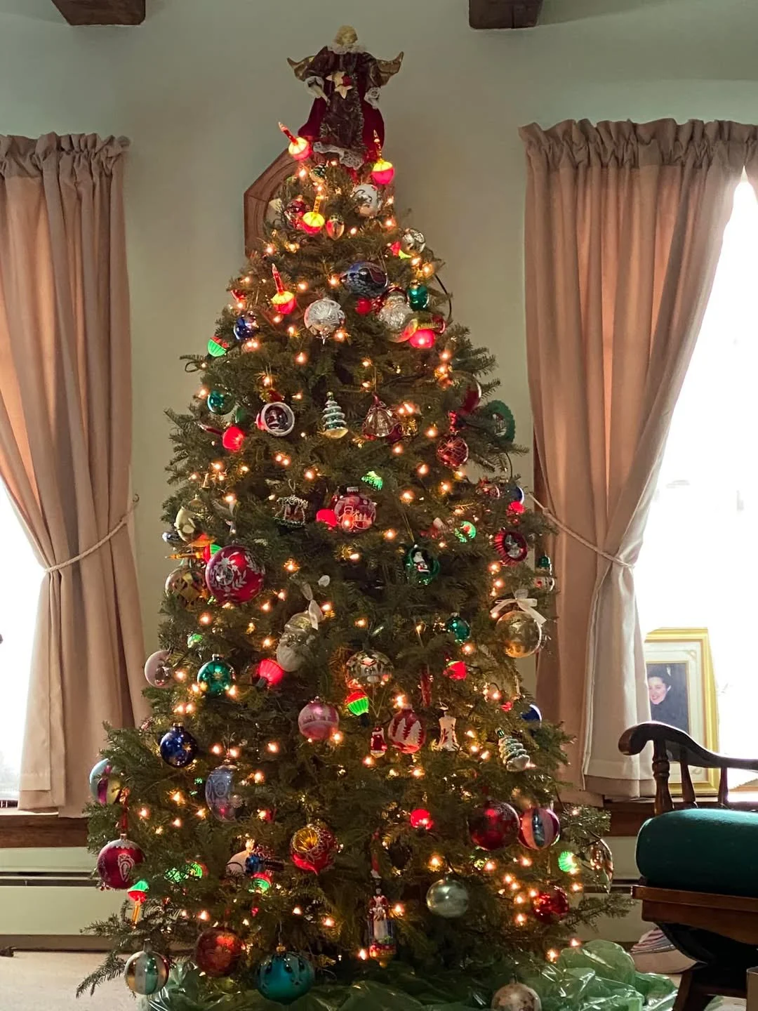 Christmas tree decorated with colorful ornaments, lights, and topped with an angel figurine, standing in a room with beige curtains and a framed photo on a side table.