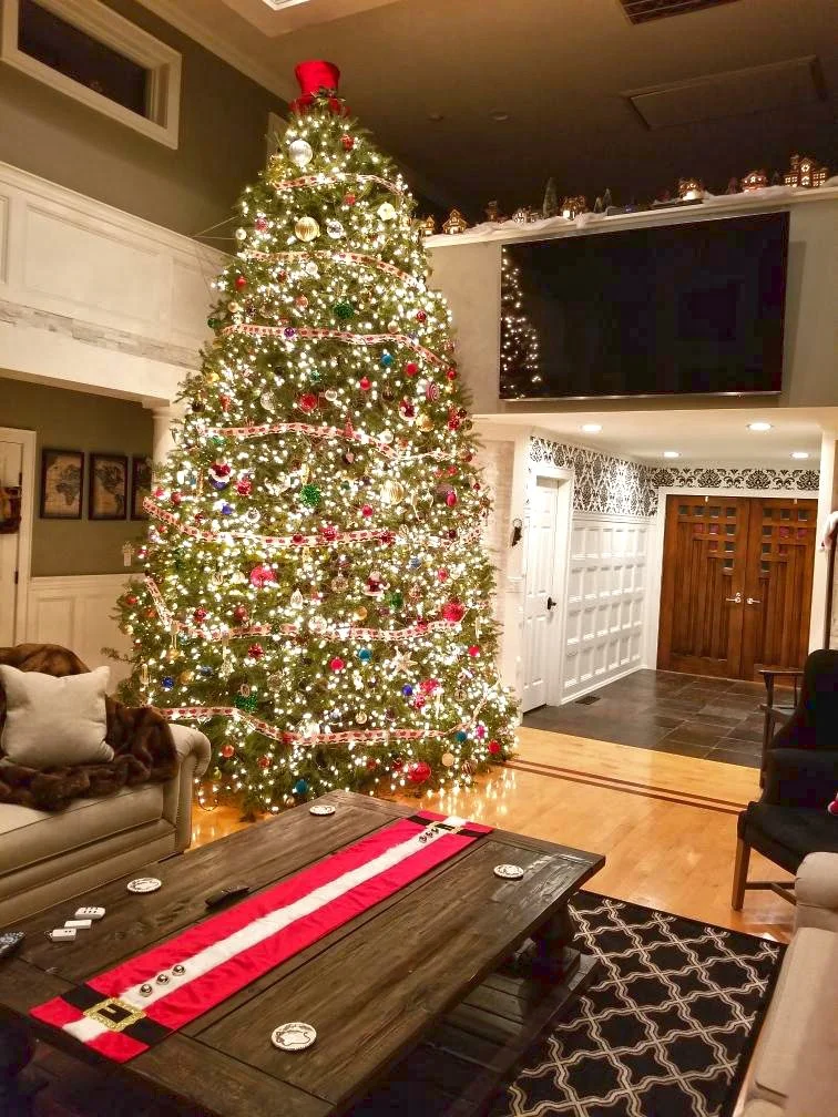 A decorated Christmas tree with lights and ornaments in a living room, with a sofa and chairs nearby, and a festive table runner on a wooden coffee table.