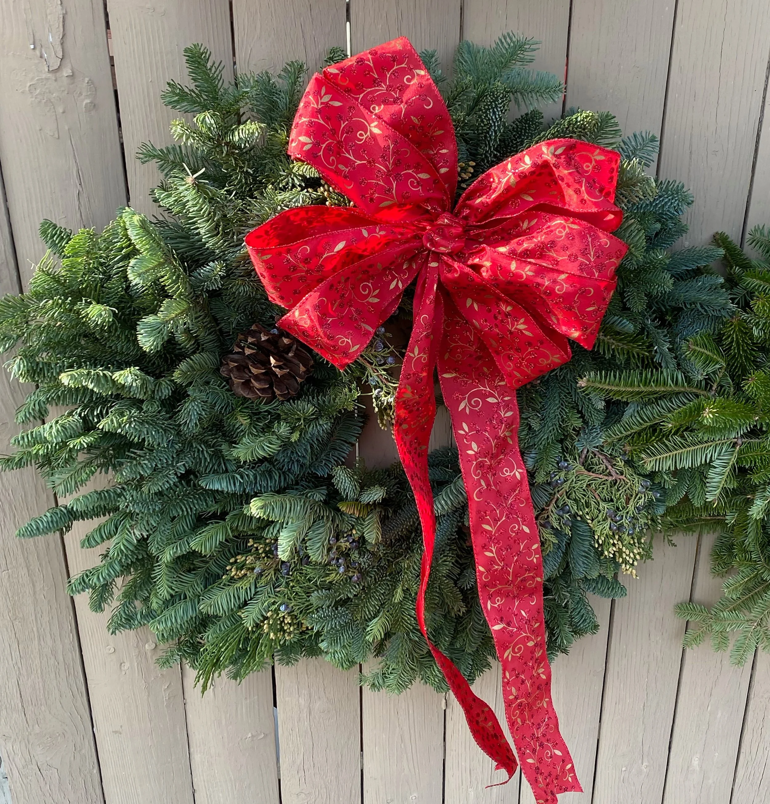 A green Christmas wreath decorated with a large red ribbon with gold patterns, hanging on a wooden exterior wall.