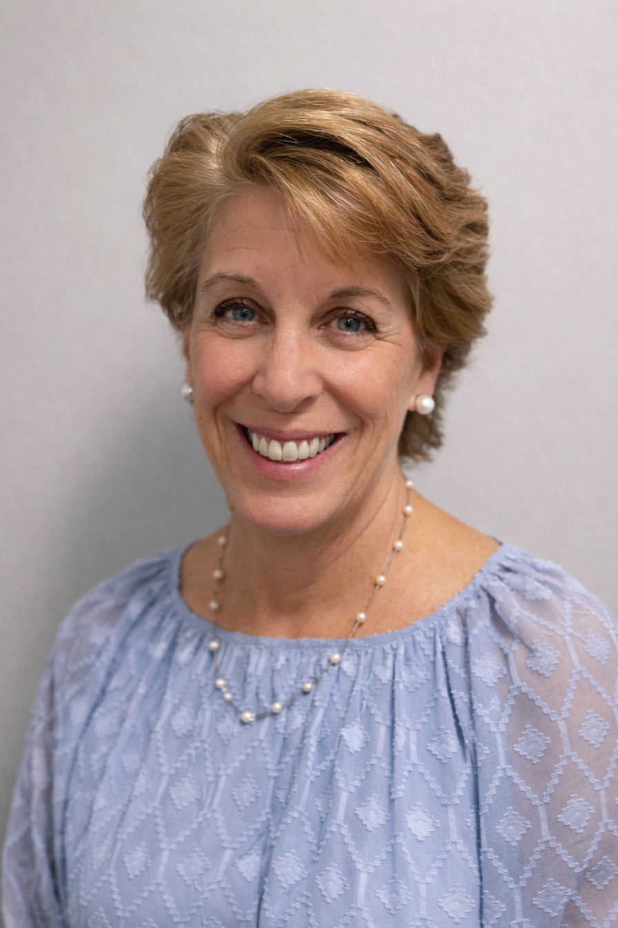 Close-up portrait of a smiling middle-aged woman with short, light brown hair, wearing a black blazer, white top, and layered necklaces with small beads. She is also wearing gold hoop earrings. The background is plain and gray.