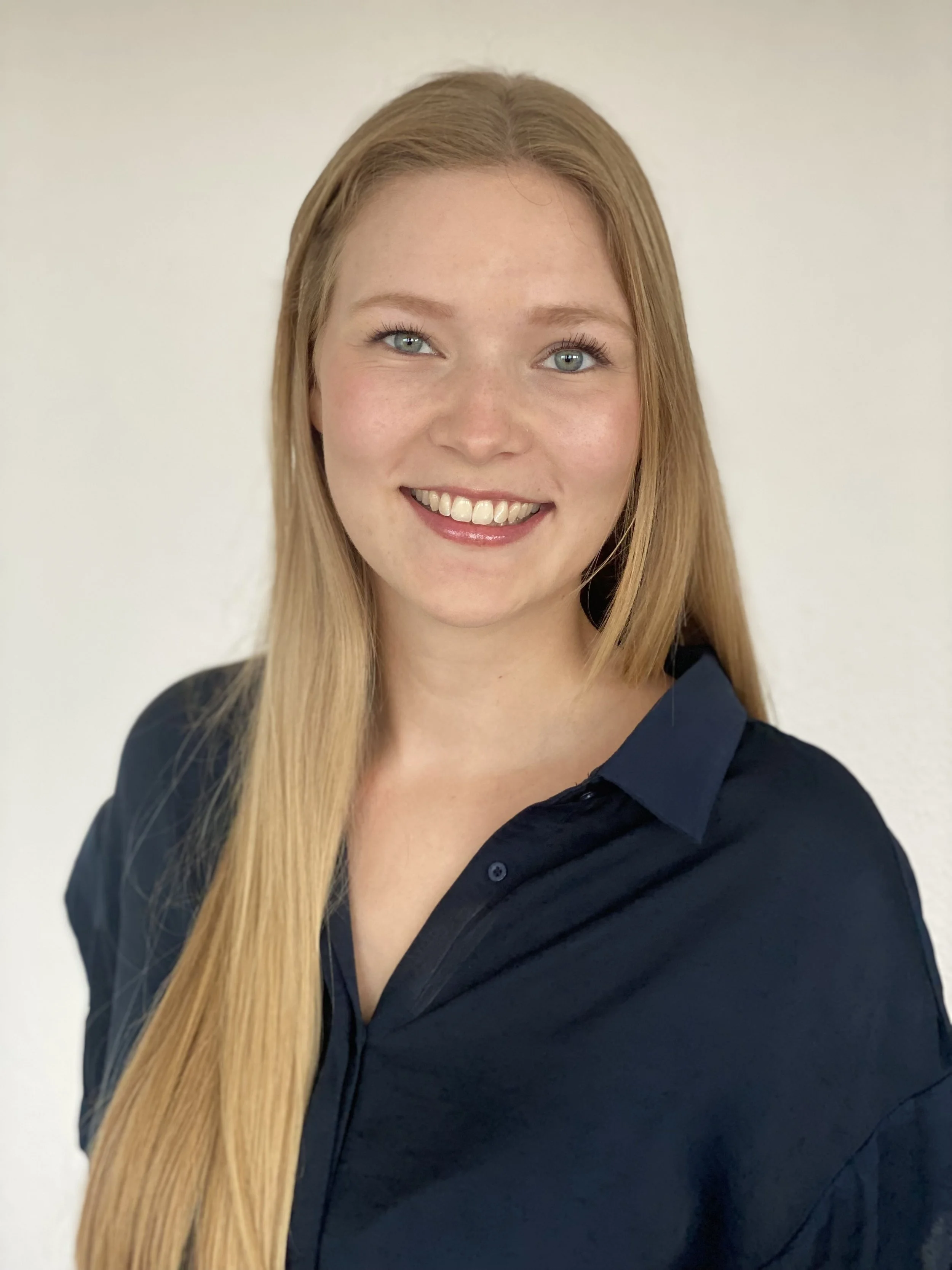 Headshot of a young woman therapist with long blonde hair, blue eyes, freckles, and a wide smile, wearing a dark navy blue collared shirt, against a plain white background.