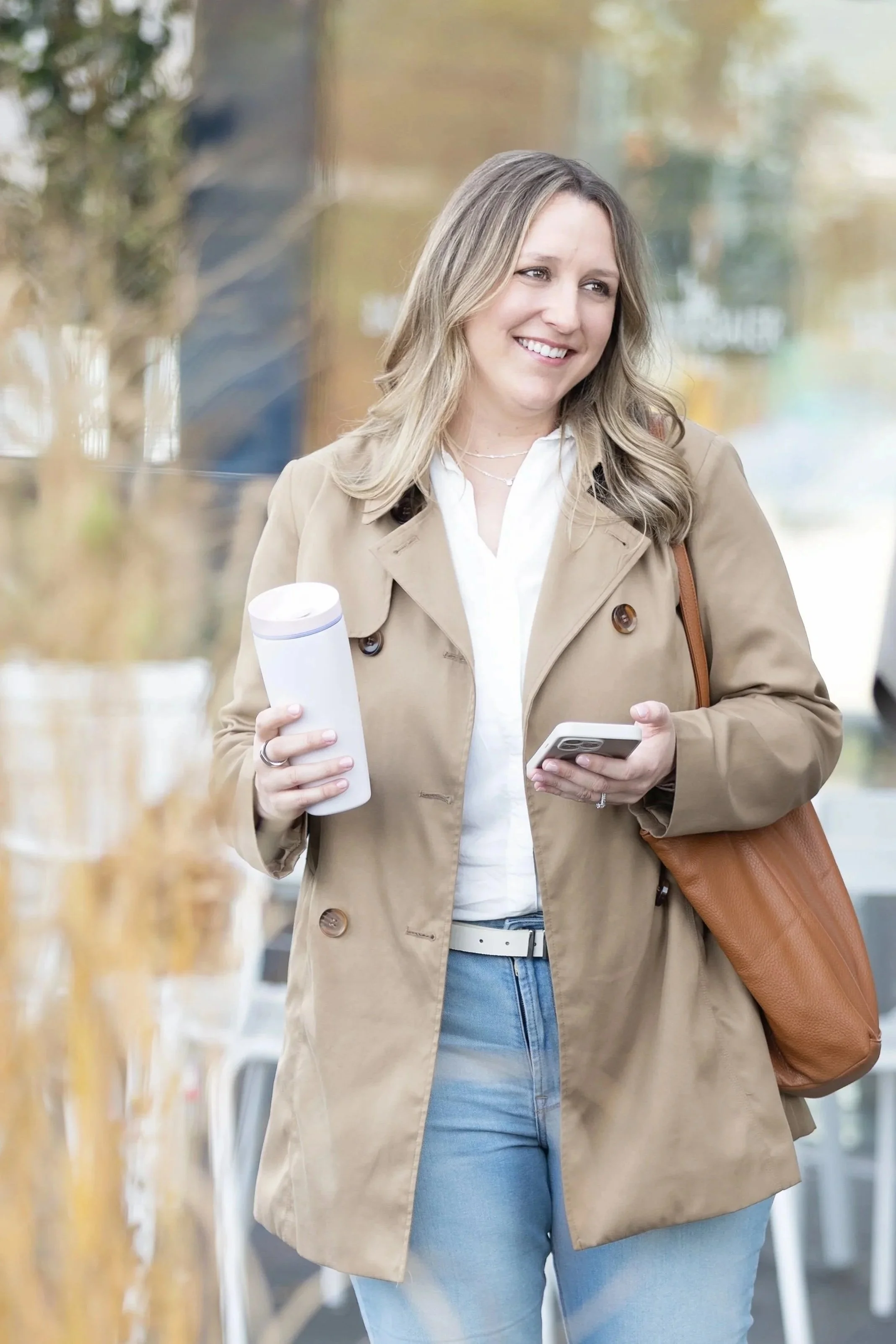 A woman with blonde hair wearing a beige trench coat, white shirt, and blue jeans, holding a white tumbler in one hand and a smartphone in the other, smiling and walking outdoors.