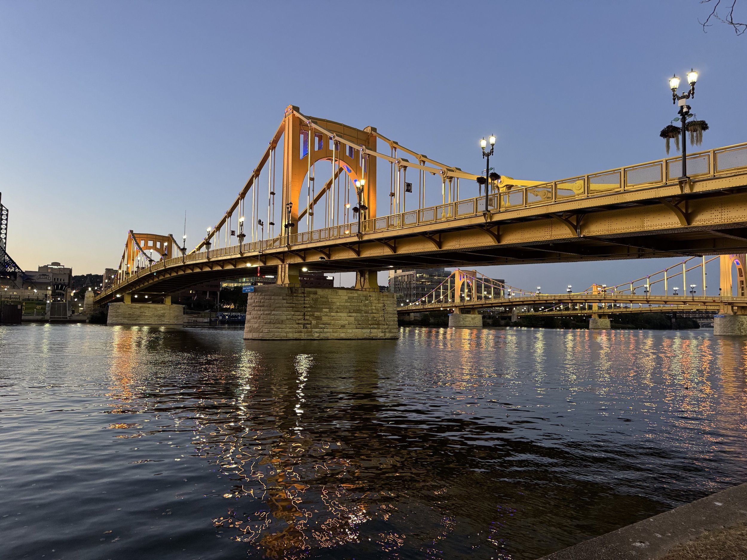 The Clarendon Bridge, a yellow arch bridge, spanning over a waterway at sunset. City lights and buildings are visible in the background.