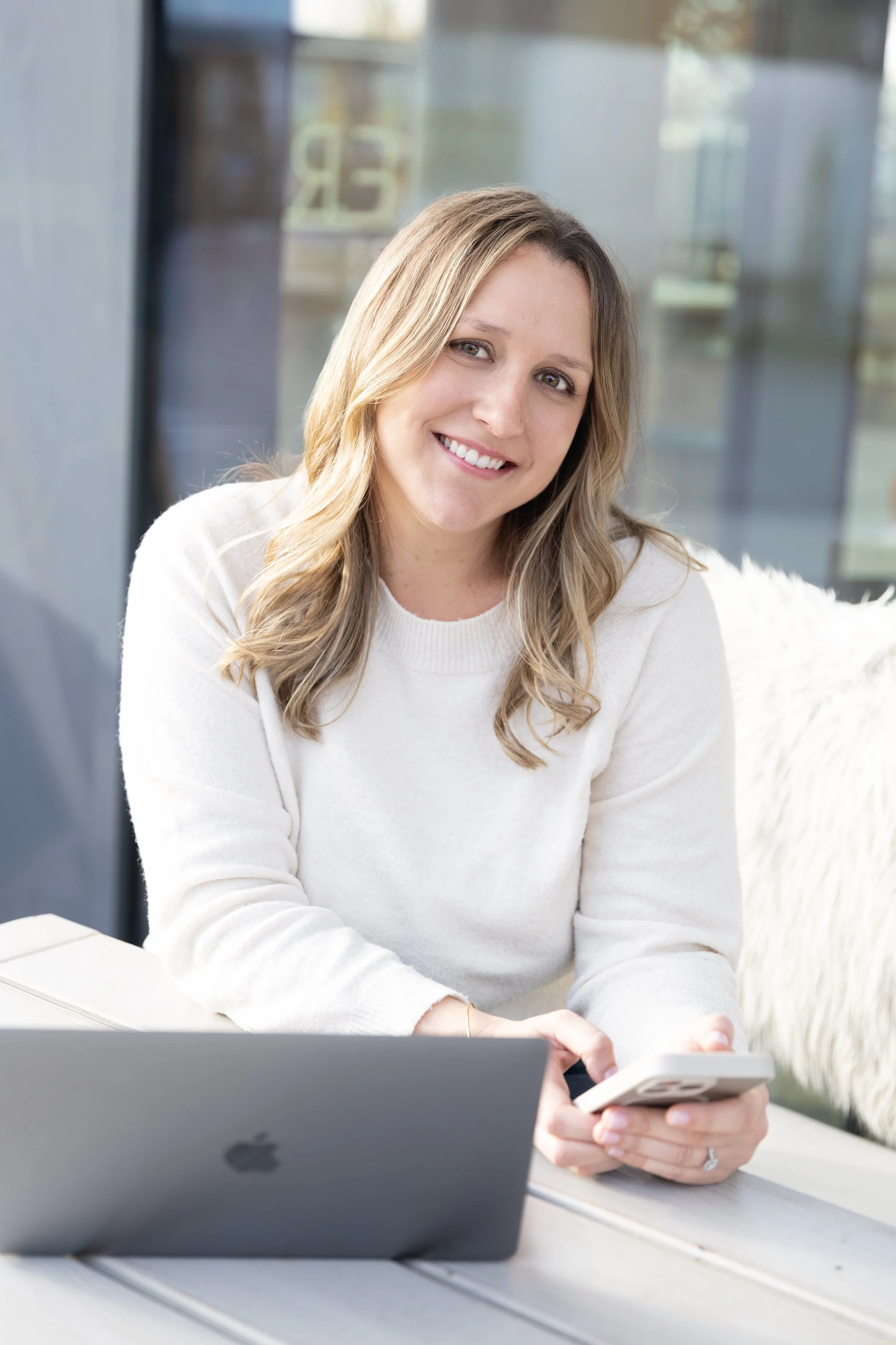 A woman sitting at a table with a laptop and smartphone, smiling at the camera, indoors with large windows in the background.
