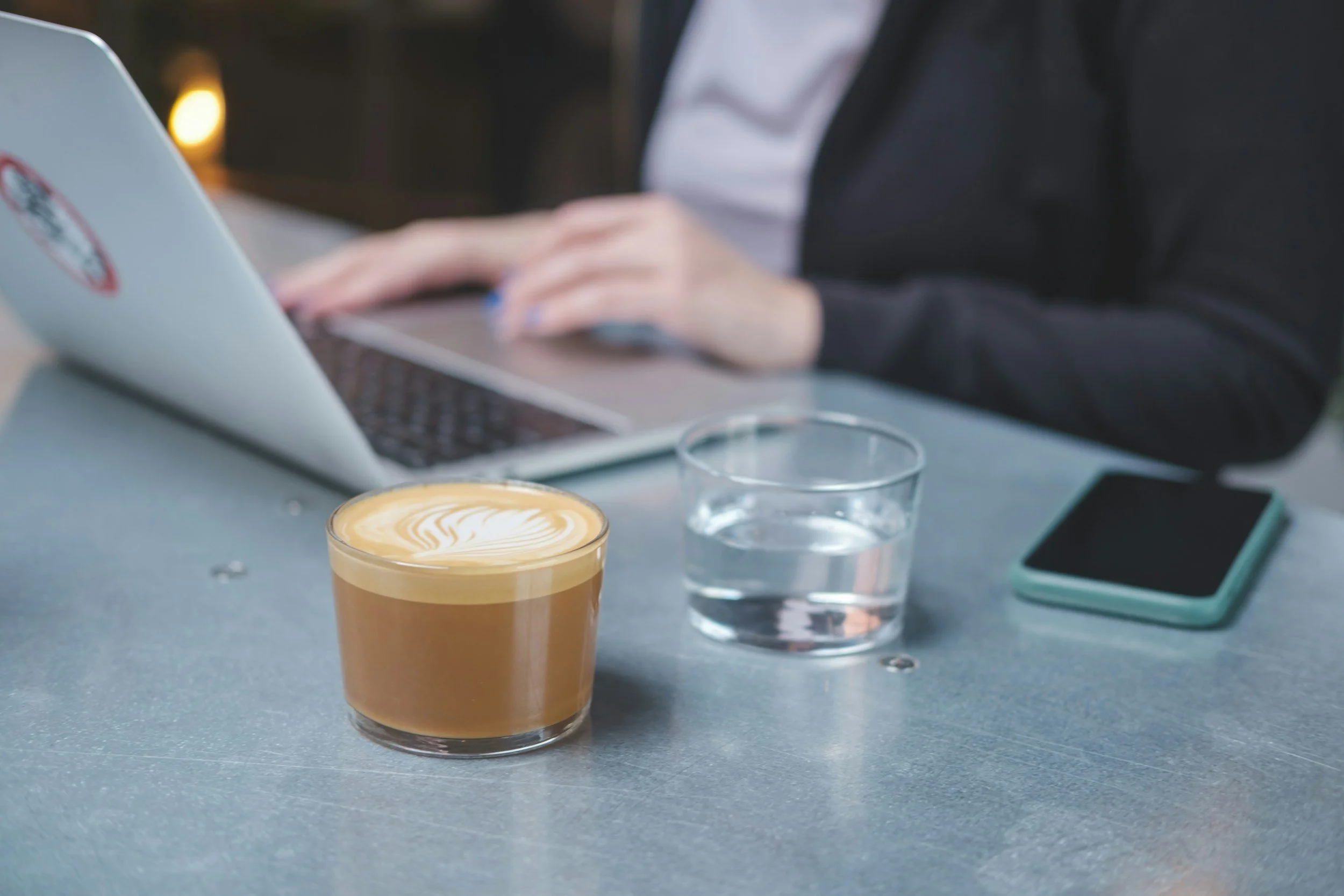 A latte in a glass with a foam design, a glass of water, a smartphone, and a person typing on a laptop at a table.