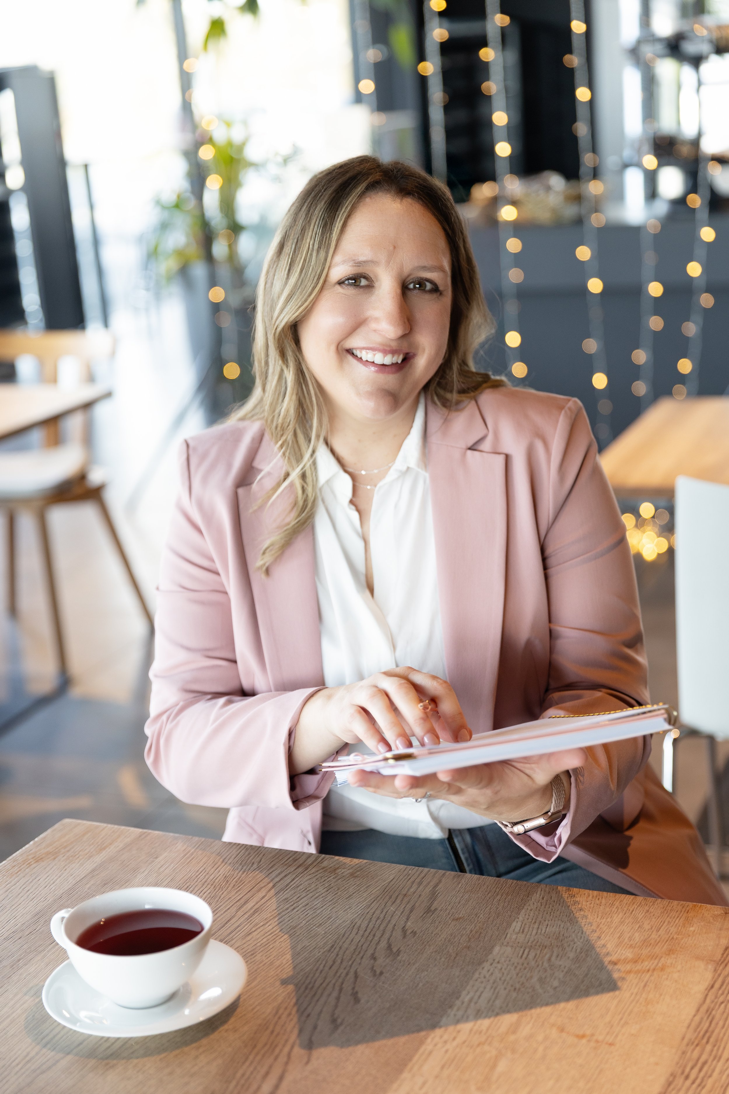 A woman with wavy blonde hair, wearing a light pink blazer over a white shirt, sitting at a wooden table in a cozy cafe. She is smiling and looking at the camera, holding a notepad or planner. There is a cup of tea on the table in front of her, and the background is decorated with string lights and greenery.