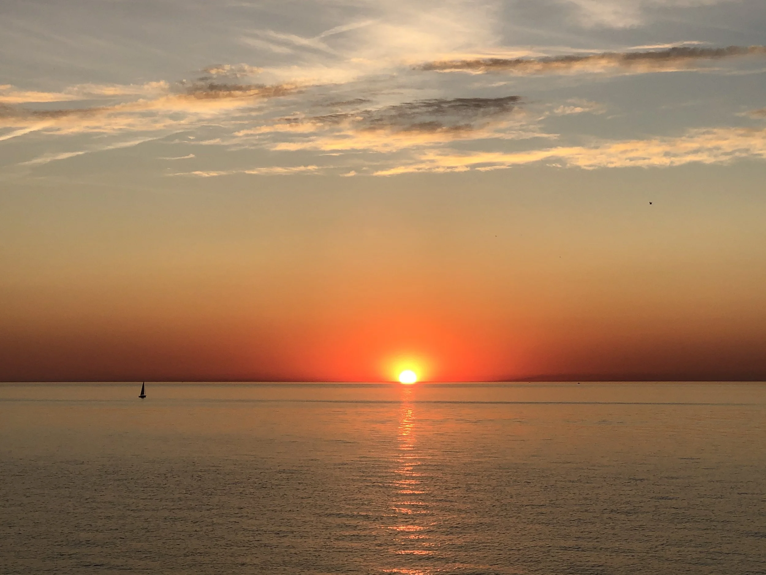 Sunset over the ocean with a sailboat on the left and colorful clouds in the sky.