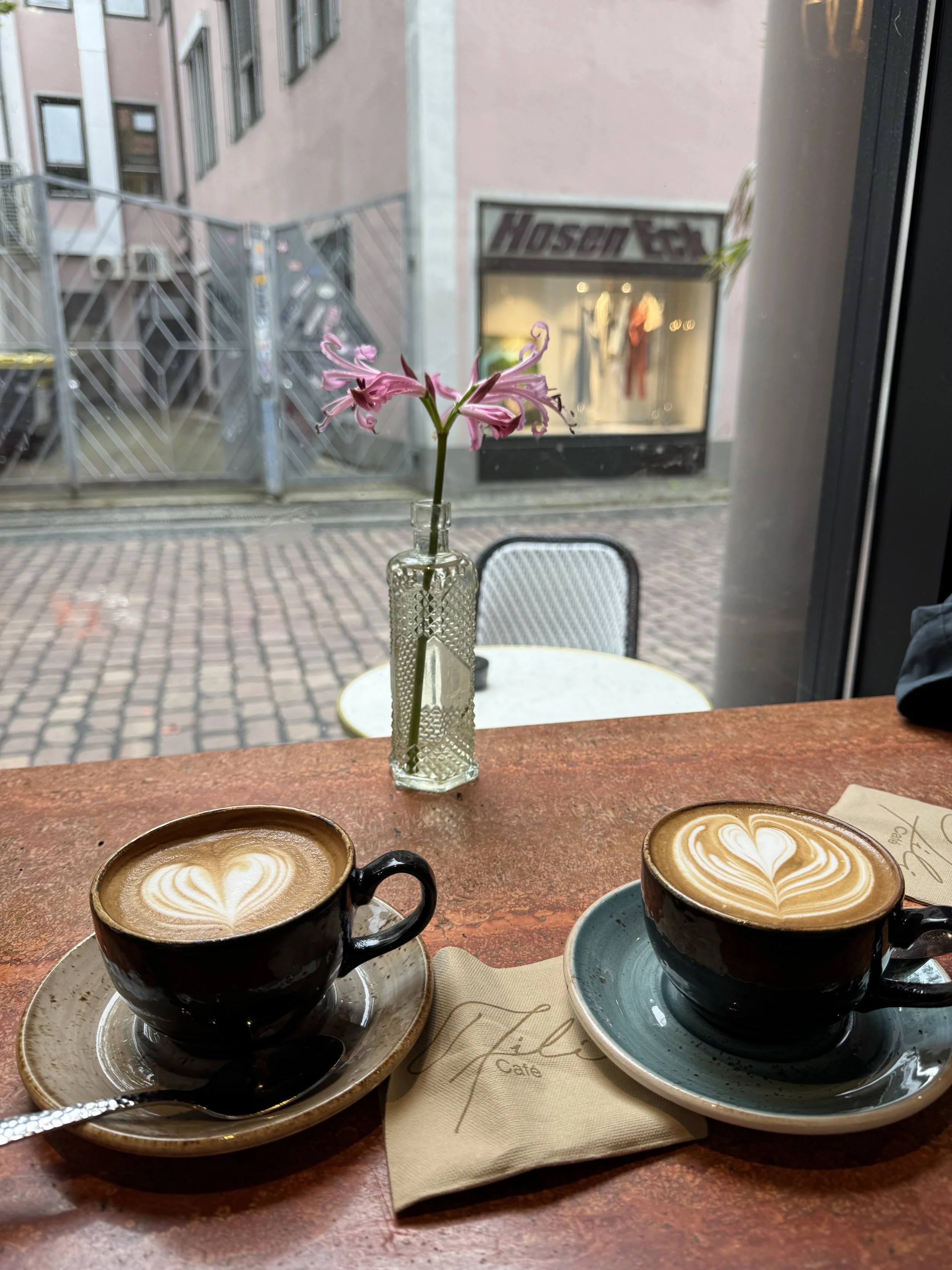 Two cups of coffee with heart-shaped latte art on a wooden table, with a small glass vase with pink flowers between them, in a cafe with a window view of a brick patio and a building with a store sign.