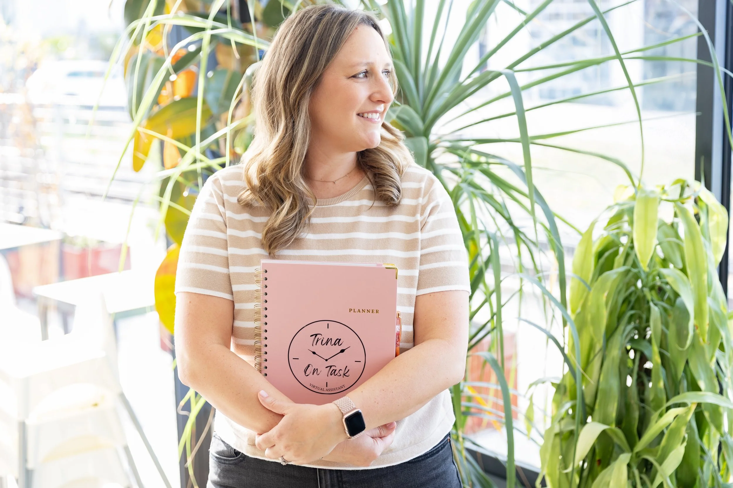 A woman standing indoors near large green plants, holding a pink planner with the words "Trina On Task" on the cover, and smiling while looking to her right.