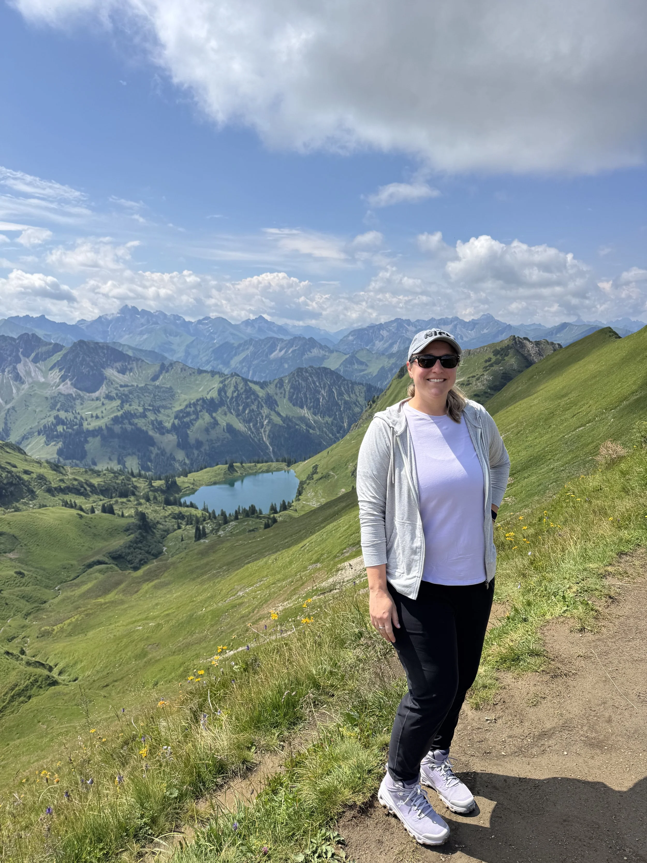 Woman wearing sunglasses, a white T-shirt, gray zip-up hoodie, black pants, and sneakers standing on a trail in a mountainous landscape with green hills, a lake, and mountain peaks under a partly cloudy sky.