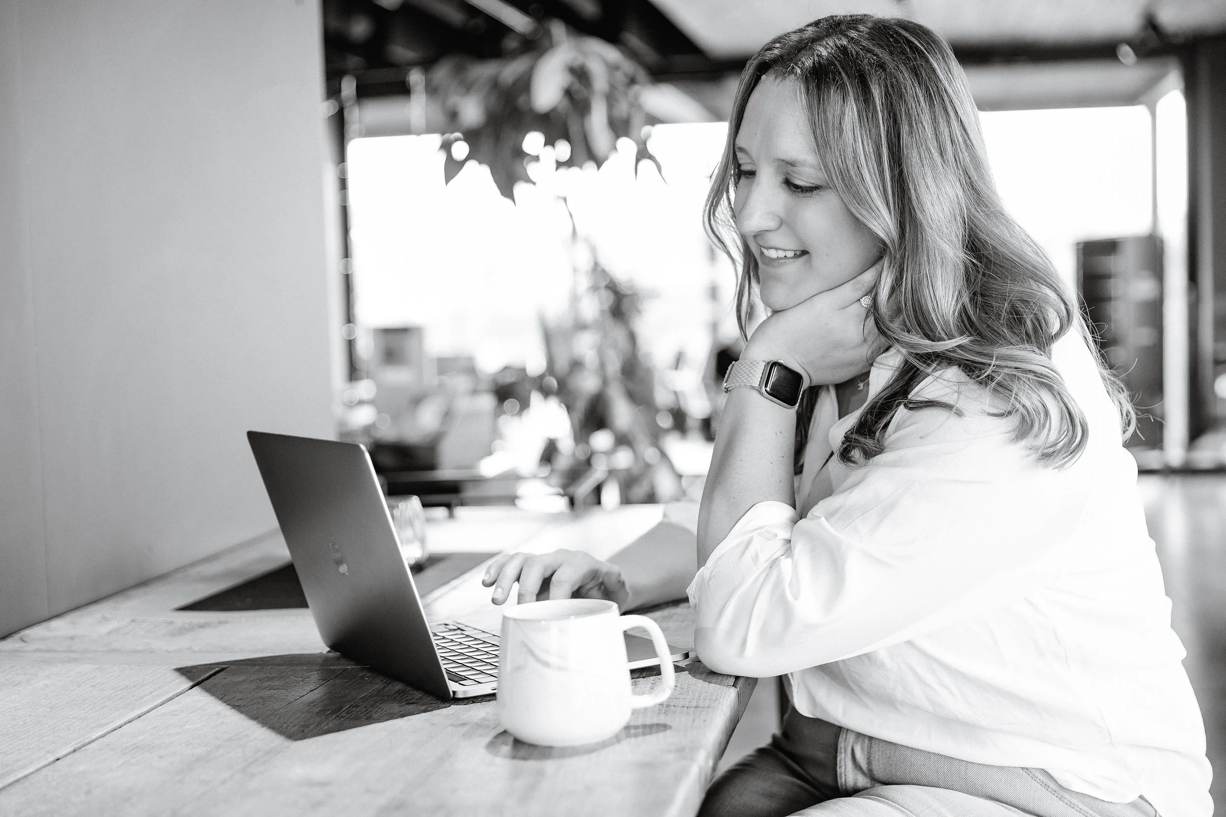 A woman with long hair, smiling, sitting at a wooden table in a cafe, looking at her laptop, with a coffee mug in front of her.