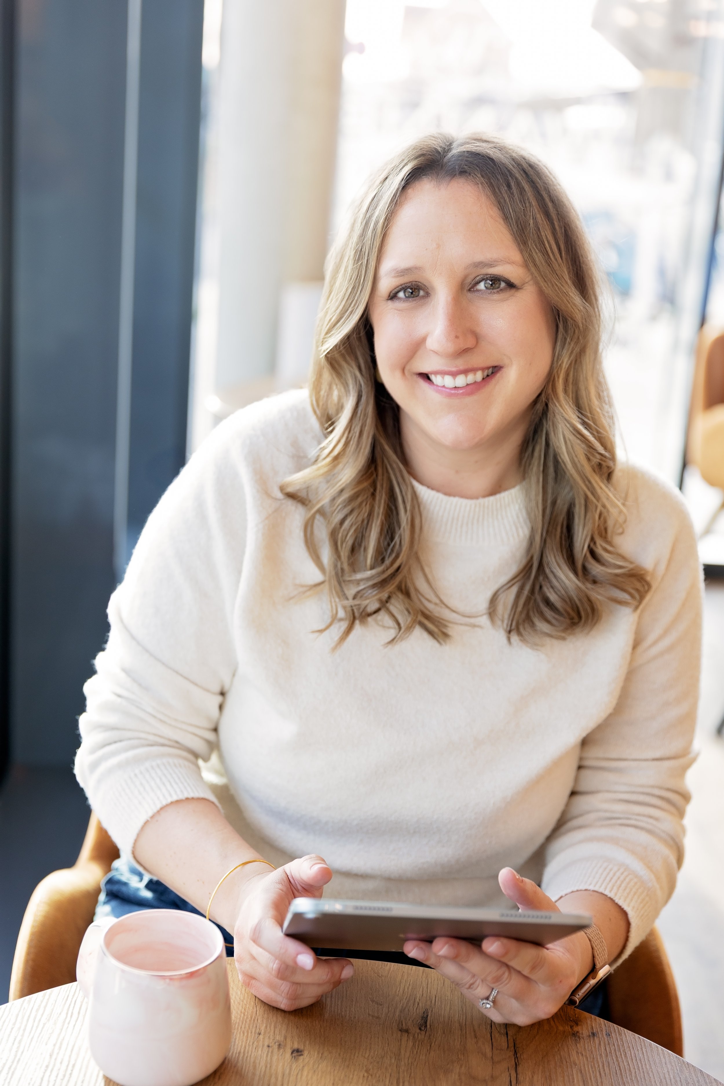 A smiling woman with wavy blonde hair wearing a cream sweater, sitting at a wooden table in a cafe, holding a tablet in both hands. A pink ceramic mug is on the table in front of her. Bright natural light is coming through the window behind her.