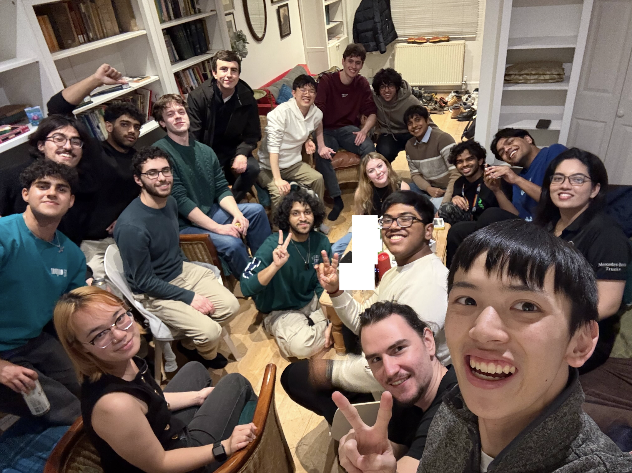 Group of young people gathered in a living room, some sitting on chairs and the floor, smiling and posing for a photo, with bookshelves and shoes in the background.
