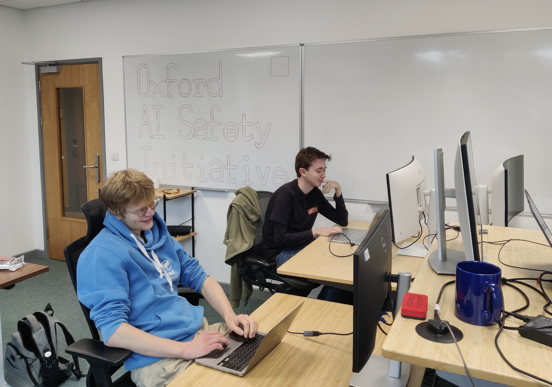 Two young men working on laptops in a classroom with a whiteboard that reads 'Oxford AI Safety Initiative'.