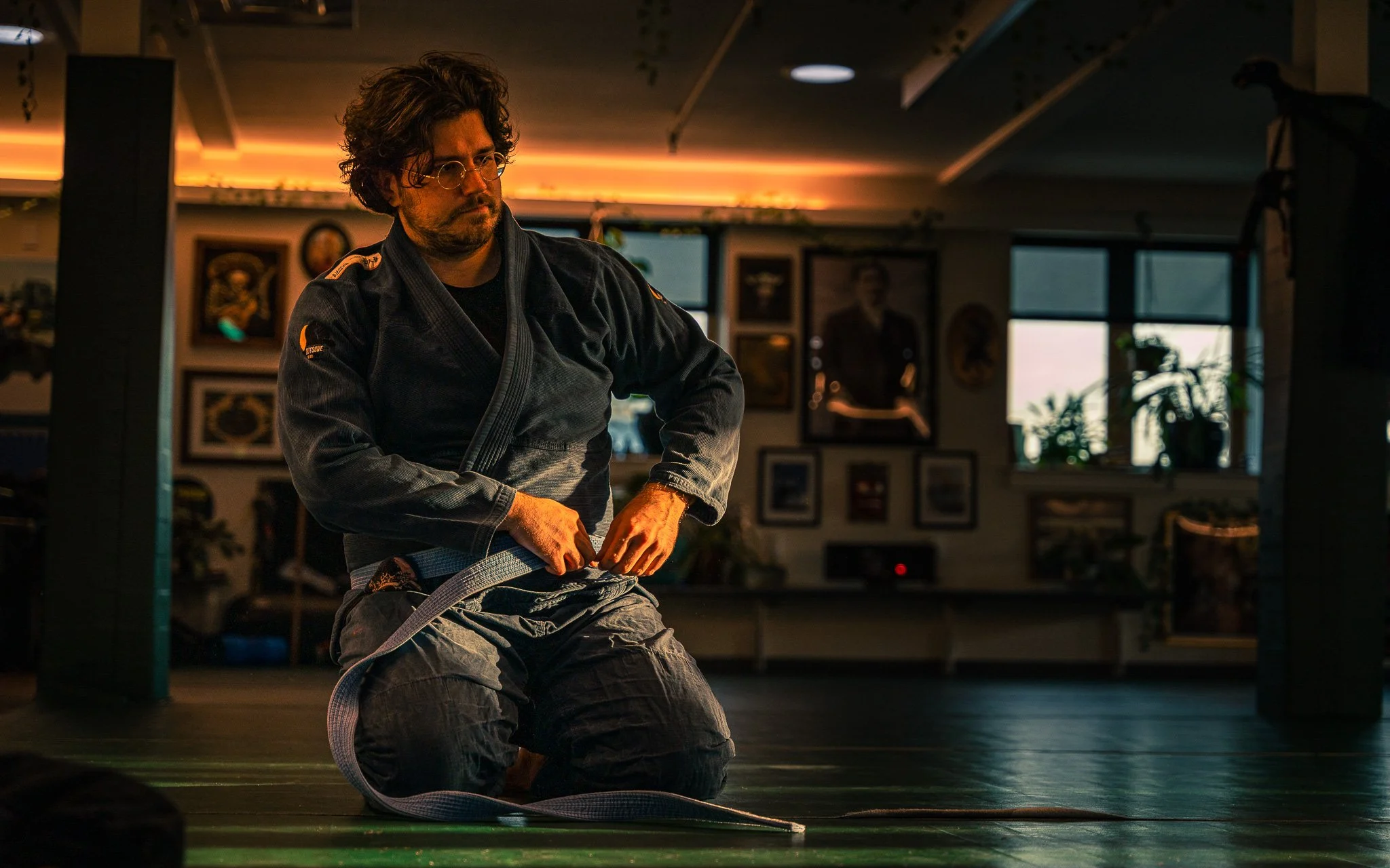 A man wearing a black Brazilian Jiu-Jitsu gi kneels on the mat, adjusting his grey belt in a dojo with framed pictures on the wall and windows in the background.