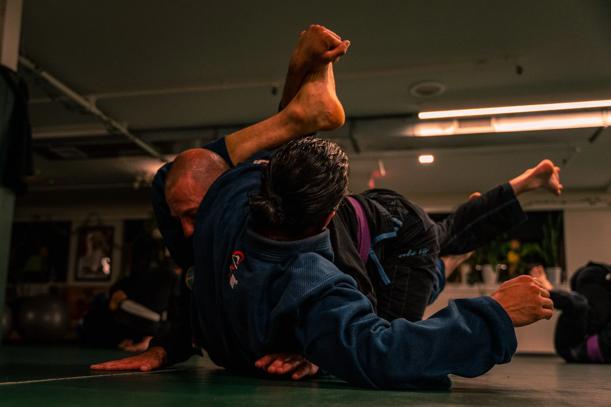 Two people practicing Brazilian Jiu-Jitsu on a mat, one in a dominant position, with their arms around the other's neck and head, while the other person attempts to escape.