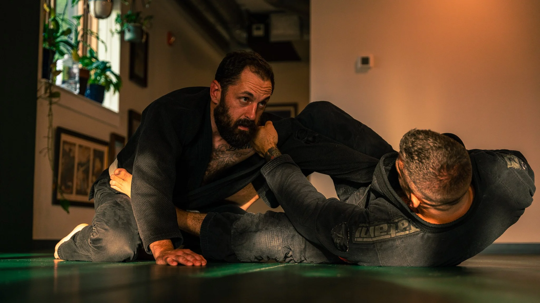 A man with a beard on top of another man on a mat, both wearing black gi uniforms, engaged in Brazilian Jiu-Jitsu or grappling, indoors.