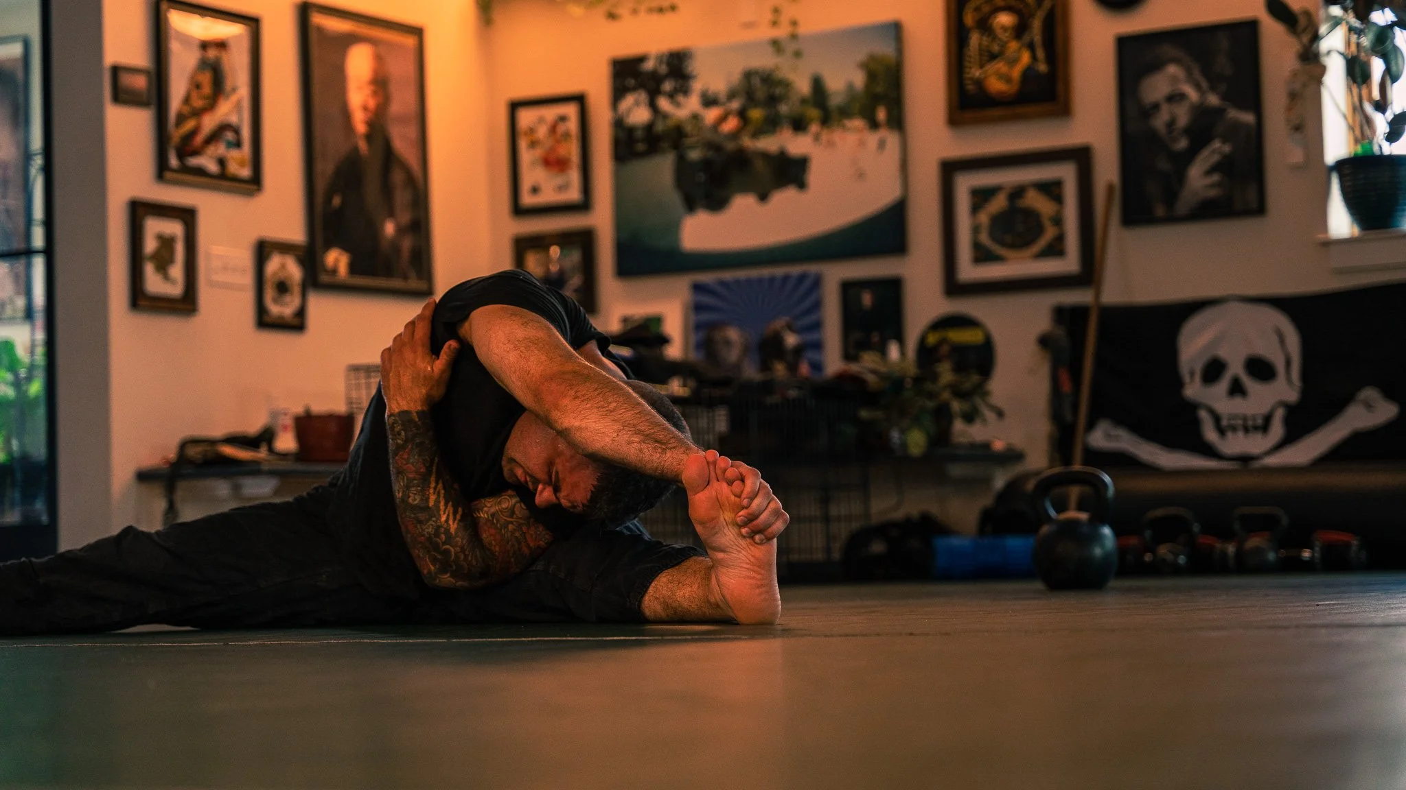 Person lying on the floor in a stretching or yoga pose, in a room with posters and artwork on the walls, and kettlebells on the floor nearby.