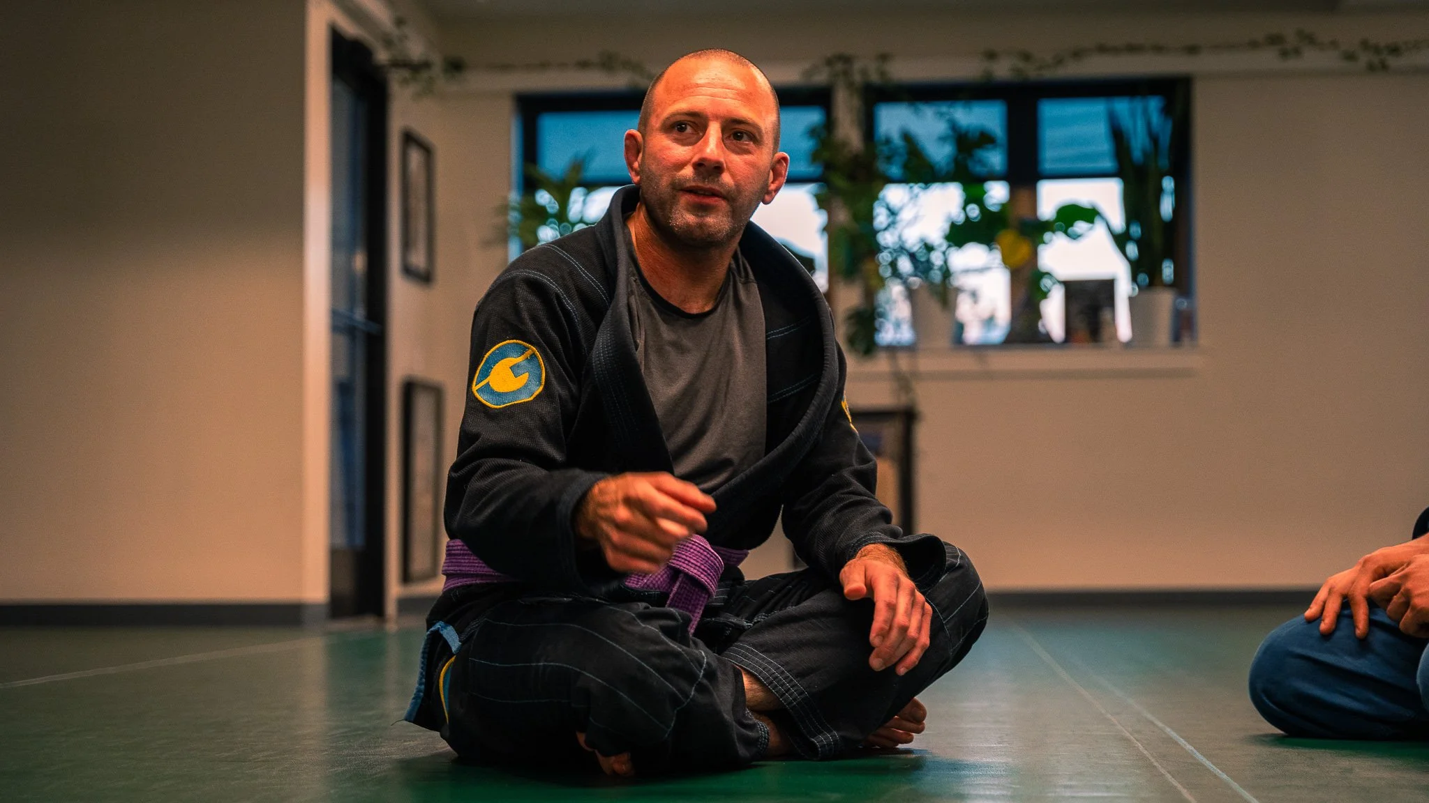 A man in martial arts attire, sitting cross-legged on the floor, speaking or explaining a technique inside a dojo with plants and windows in the background.