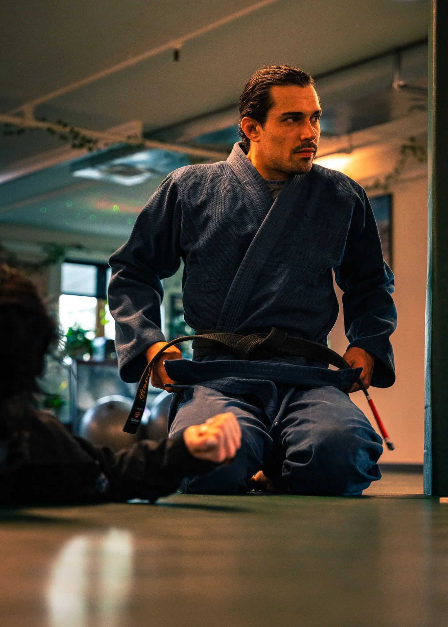 A man wearing a blue martial arts gi kneels on a mat during a jiu-jitsu training session, gripping the belt of a opponent lying on the floor.