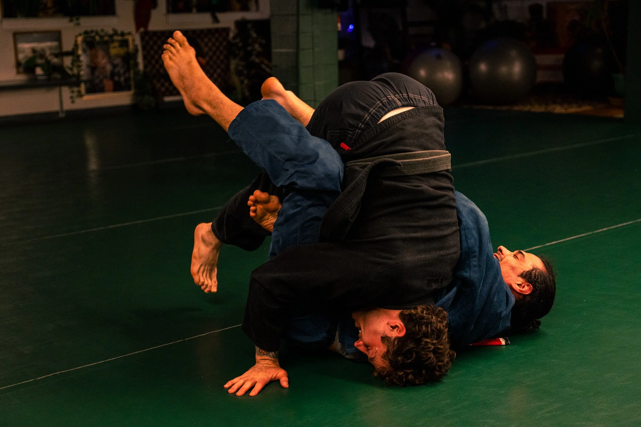 Two people practicing Brazilian Jiu-Jitsu on a green mat, with one person in a black gi on top and the other person on the bottom in a blue gi, in a gym with exercise balls in the background.