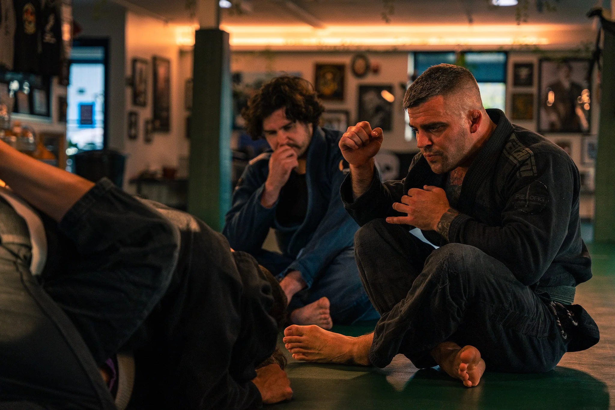 Two men practicing Brazilian Jiu-Jitsu on a mat in a gym, with framed pictures on the walls and a green support beam in the background.