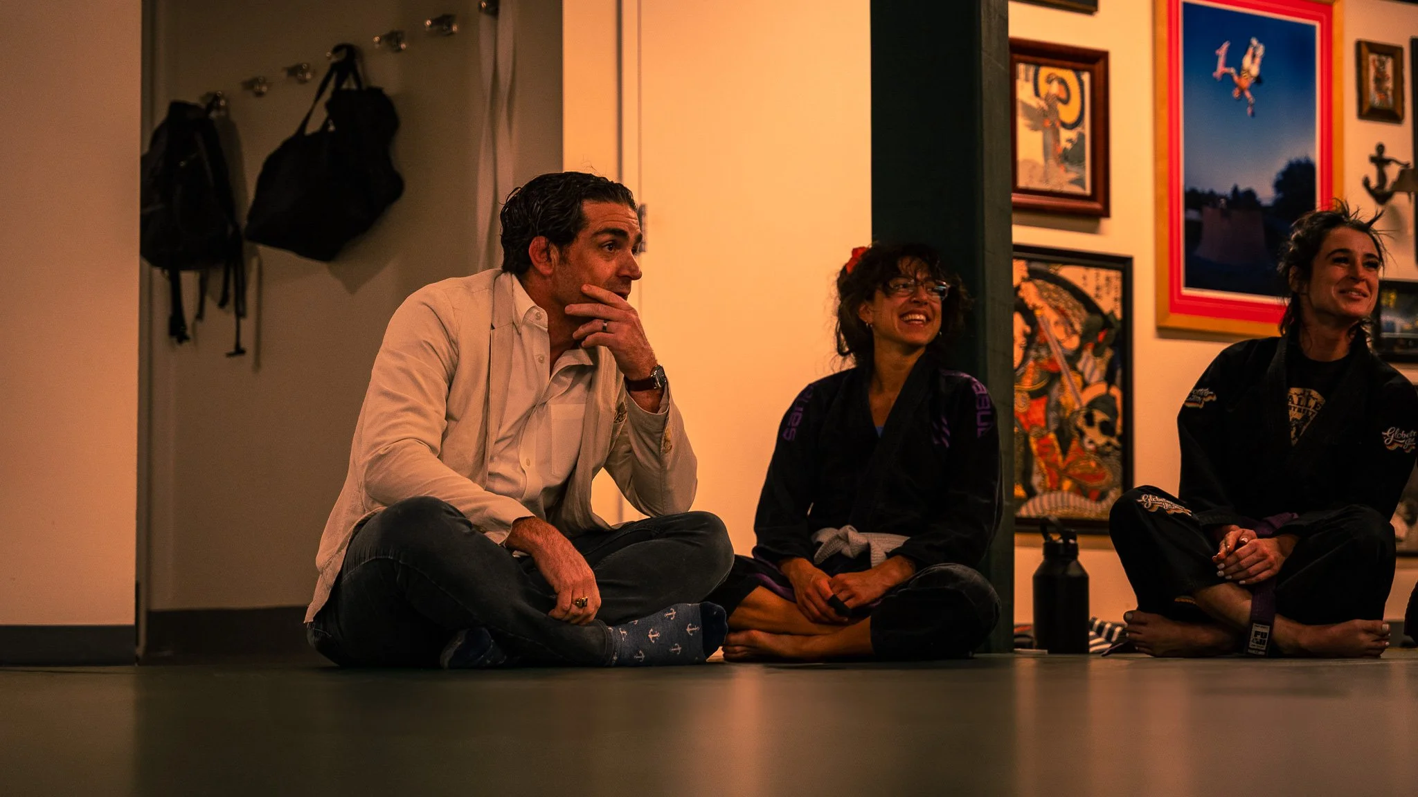 Three people sitting on the floor in a room, two women in martial arts uniforms and a man in casual clothes, smiling and engaging in conversation, with wall art and backpacks in the background.