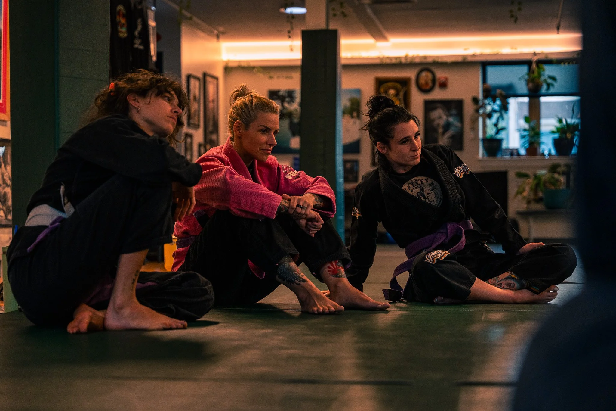 Three women sitting on the floor in a martial arts gym, listening attentively.