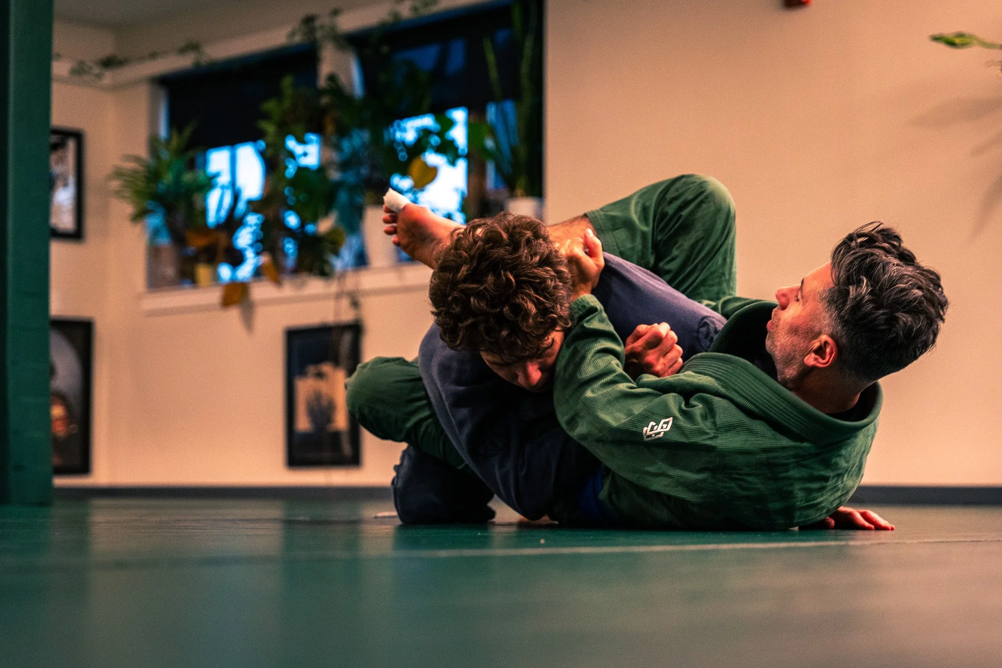 Two people practicing Brazilian Jiu-Jitsu on a mat in a gym, with one person in a green gi and the other in a dark gi, engaged in a grappling position.