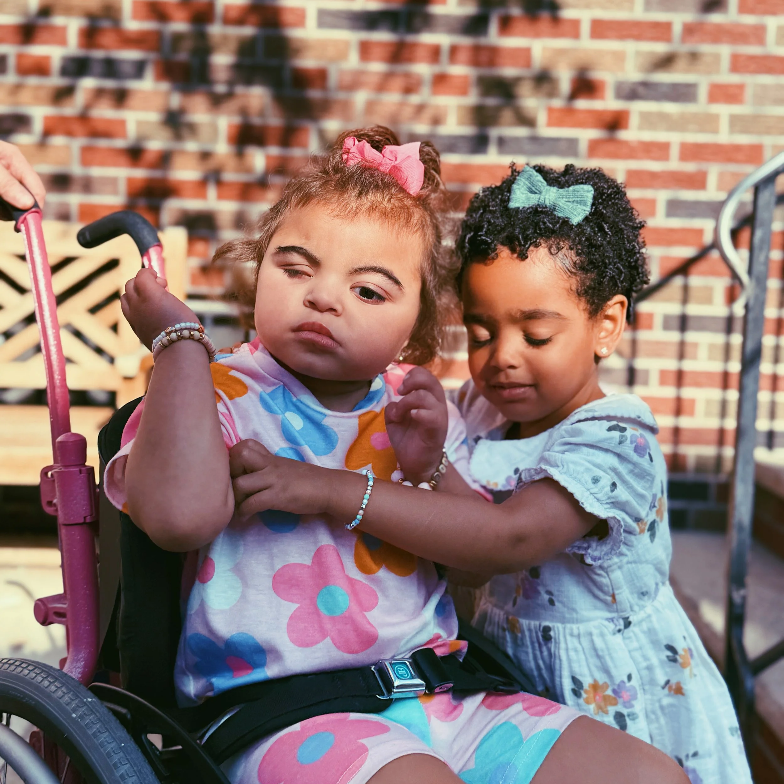 Two young girls hugging outside with a brick wall background. One girl is sitting in a wheelchair, wearing a floral dress with her hair in a pink bow, and has a serious expression. The other girl is standing next to her, with a blue bow in her curly hair, and is gently hugging her.