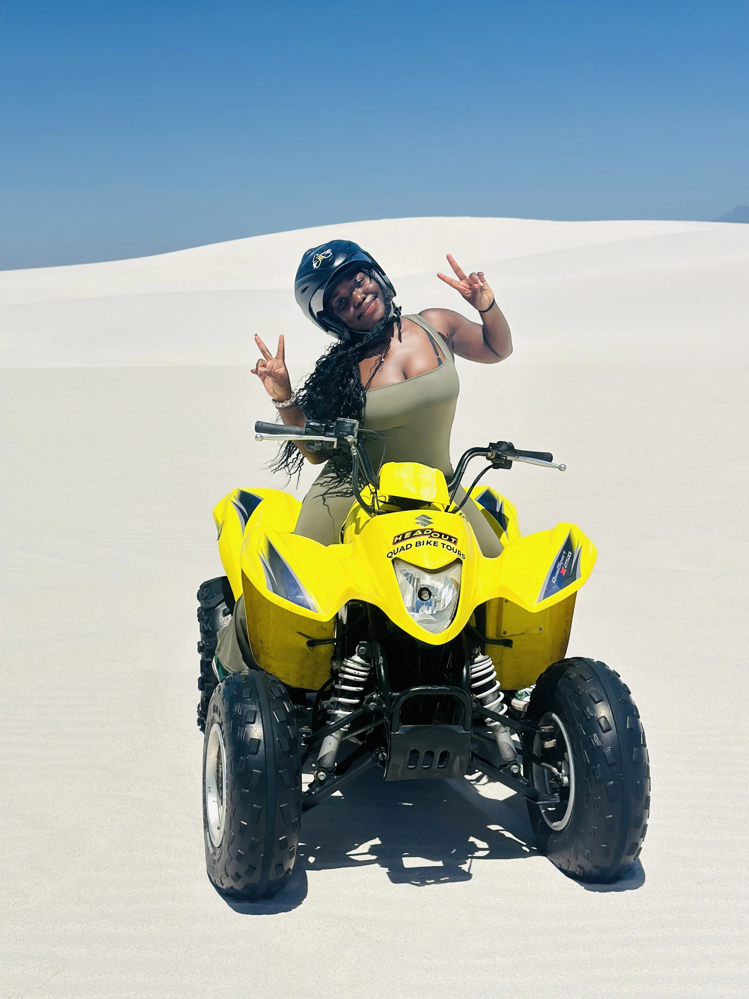 Woman in a helmet on a yellow all-terrain vehicle in a white desert with a clear blue sky, making peace signs and smiling.