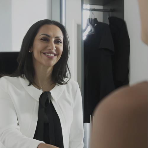Eine Frau lächelt beim Gespräch in einem Büro, im Hintergrund eine offene Garderobe mit dunkler Kleidung.