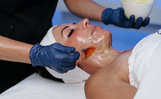 Eine Frau erhält eine Gesichtsbehandlung mit einer Gesichtsmaske, während sie auf einer Behandlungsliege liegt, und eine Kosmetikerin trägt blaue Handschuhe.