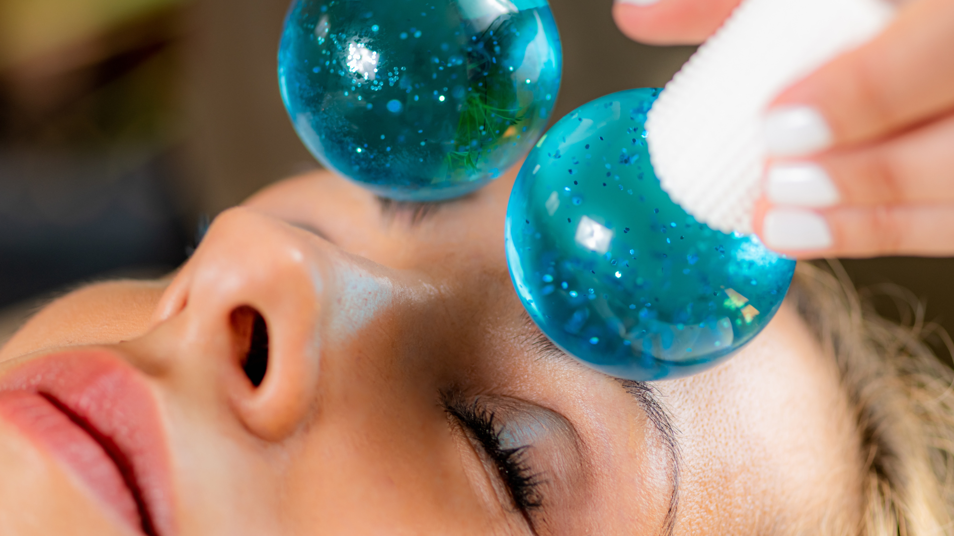 Person erhält eine Schleuse gegen die Augen mit blauen, glitzernden Wasserbälgen, während eine Hand ein Tuch auf die Lider legt.