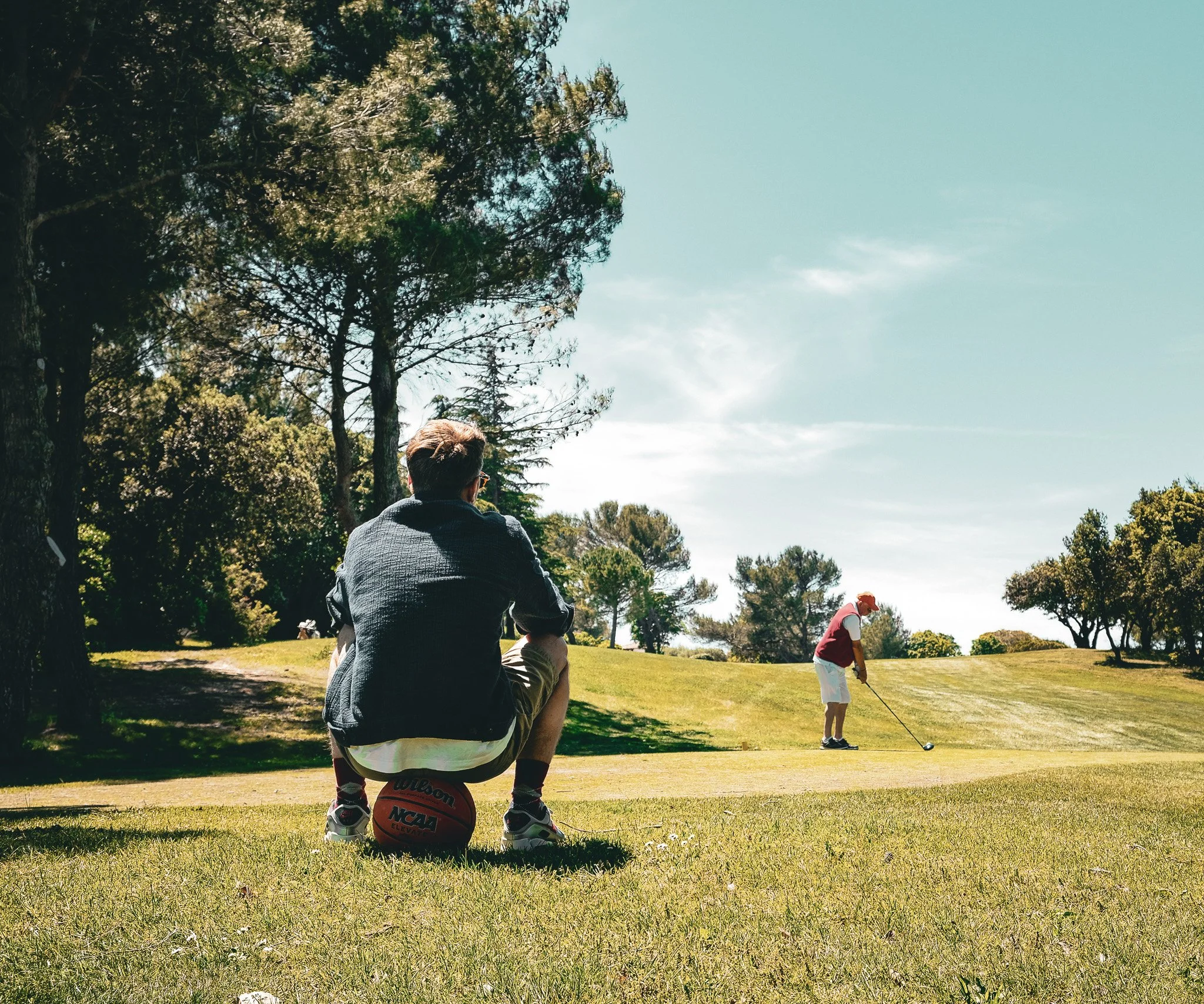 Deux personnes jouent au golf sur un terrain en plein air, parmi des arbres et un ciel bleu.
