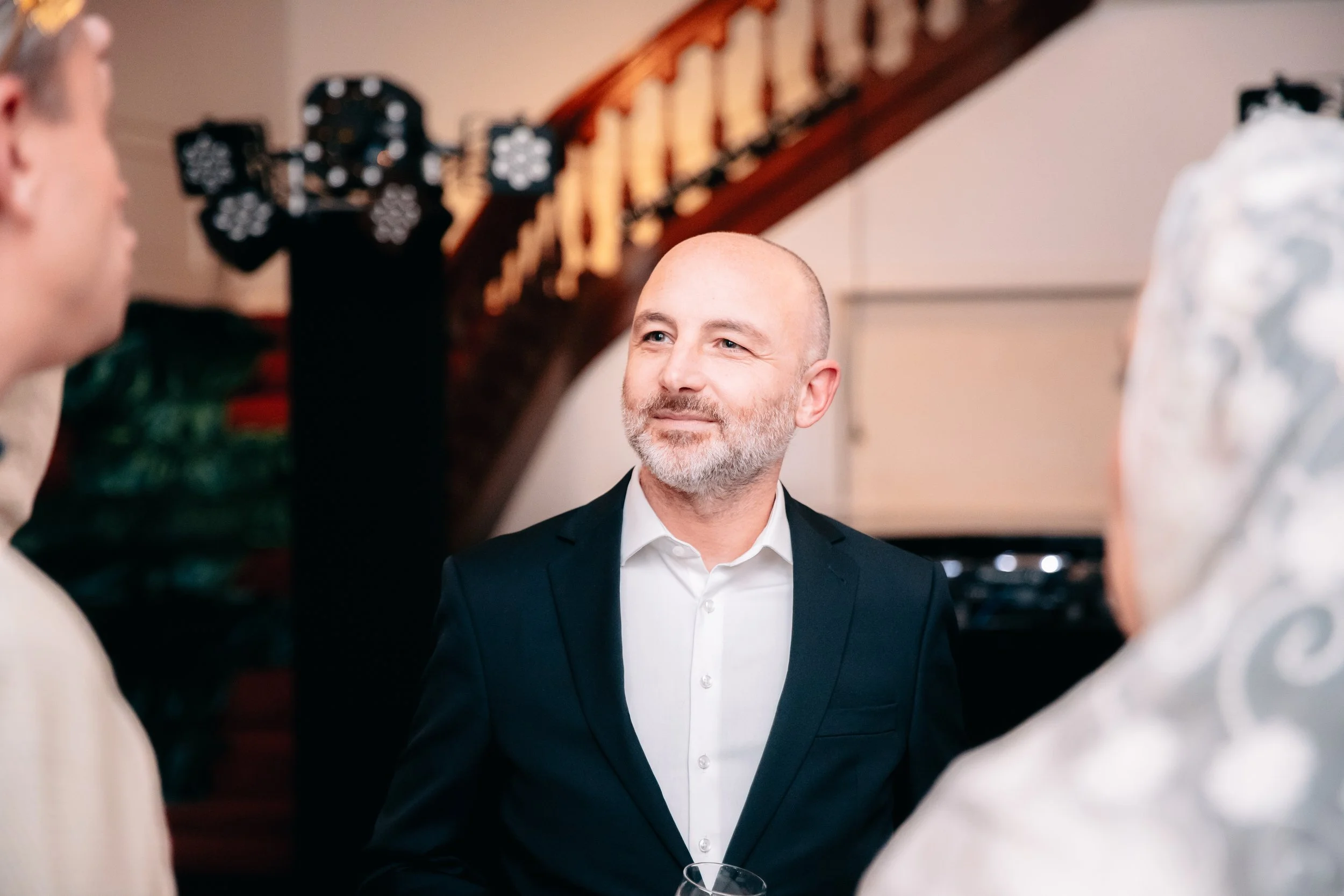 Groupe de personnes lors d'une réception, avec un homme au centre portant un costume noir et une chemise blanche, souriant légèrement. Conversation dans un espace intérieur chaleureux.