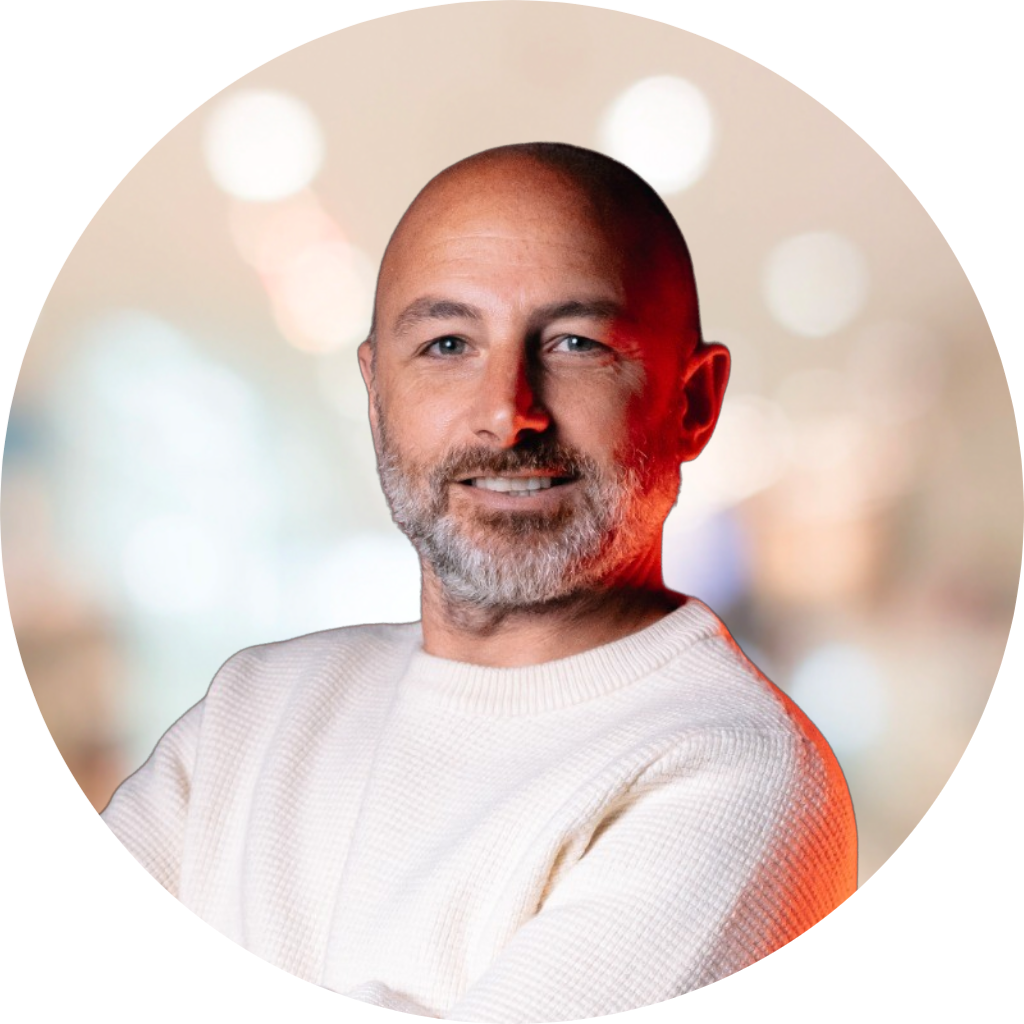 Homme souriant avec barbe, portant un pull blanc, fond flou avec des lumières