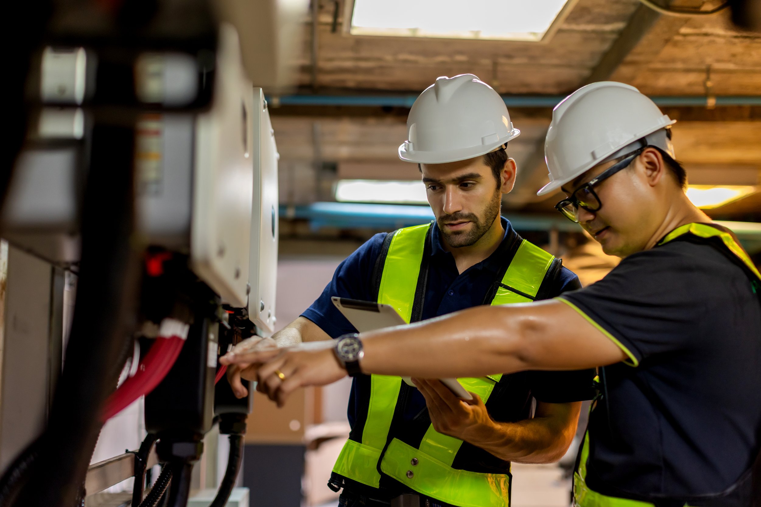Two male workers with white hard hats and safety vests working on electrical equipment in an industrial setting.