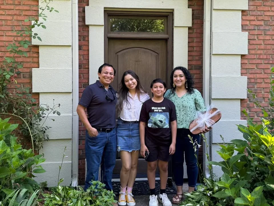 Four people standing in front of a brown front door on a brick house, smiling. Two women and two kids, with plants on either side of the doorway.
