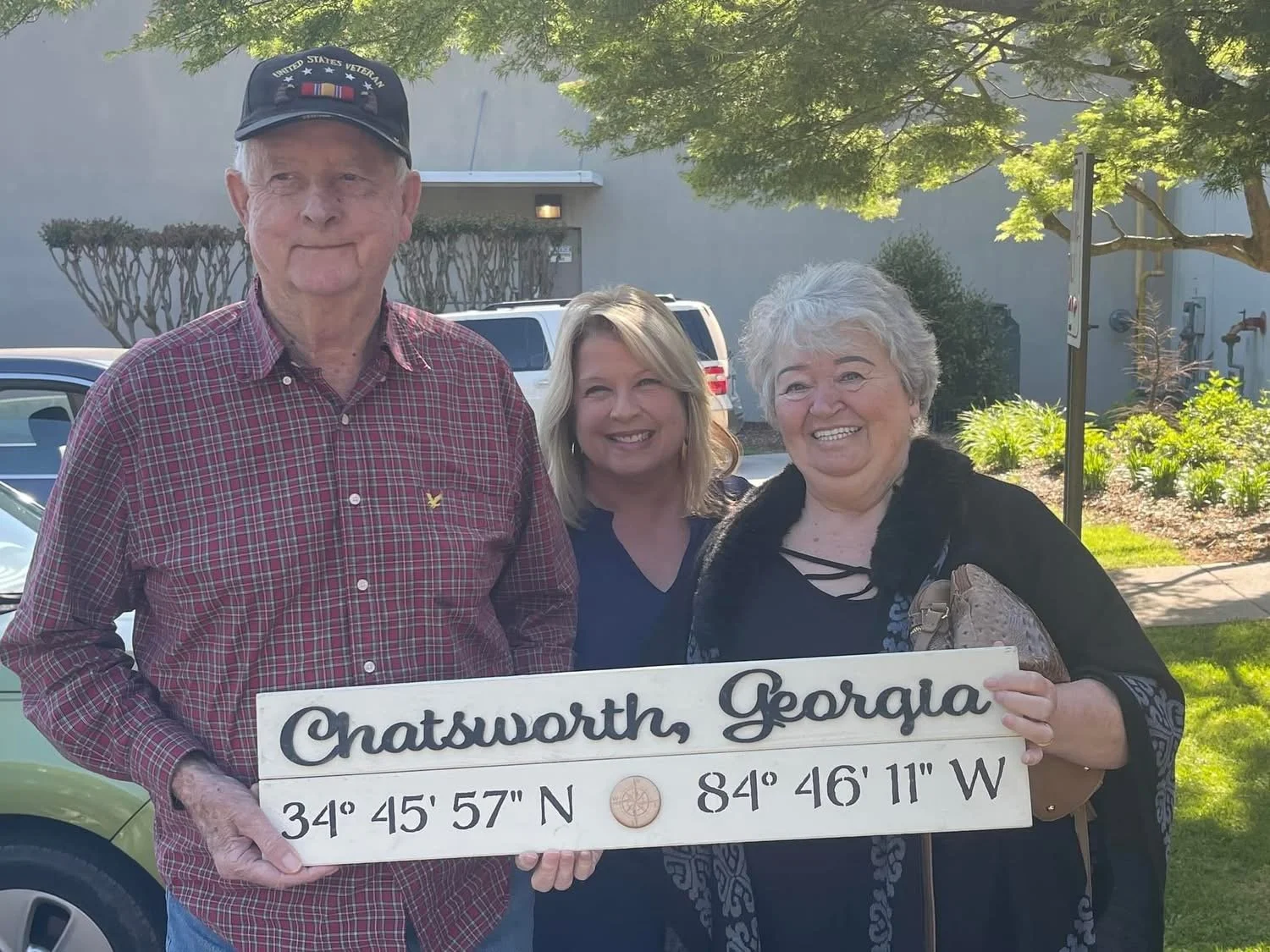 Three smiling people standing outdoors in sunlight, holding a white sign with the text 'Chatsworth, Georgia' and geographic coordinates.