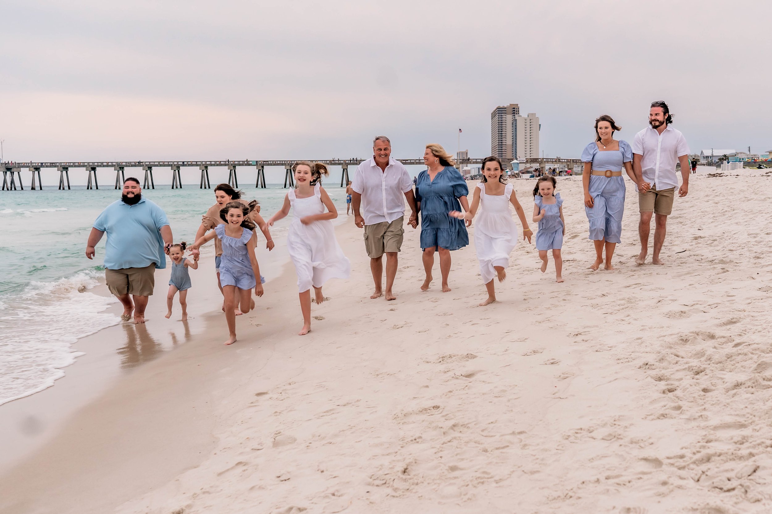 Family of eleven people, including children and adults, walking and playing on a beach with a pier in the background.